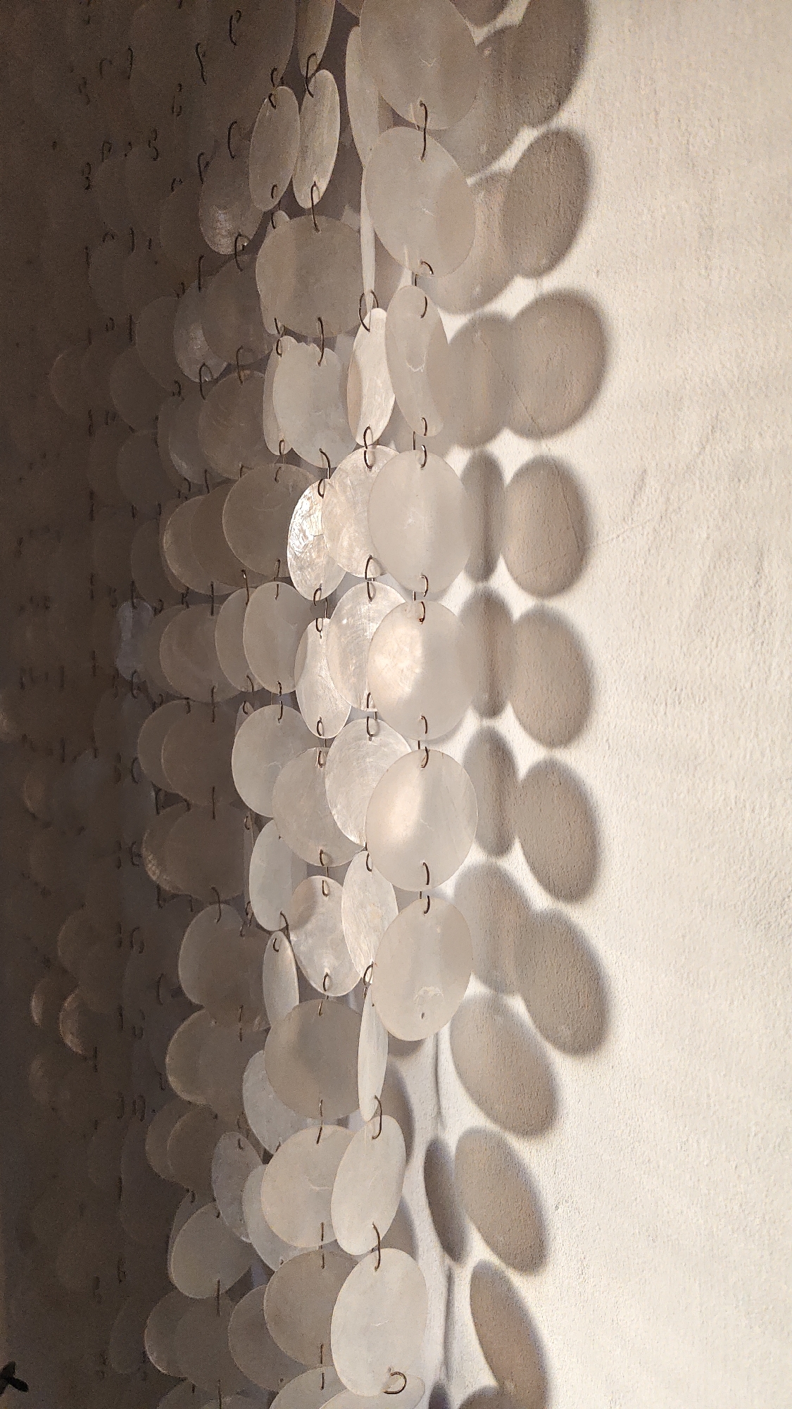 A close-up, vertical, eye-level shot shows a beaded curtain made of numerous flat, circular, mother-of-pearl discs. The discs are translucent and have a pearlescent sheen, reflecting light and creating a shimmering effect. They are strung together with small metal rings, forming multiple vertical strands that hang parallel to each other. The curtain casts distinct, oval-shaped shadows on a textured, light-colored wall to its right, indicating a light source from the left. The discs are arranged in rows, with slight variations in their spacing and orientation, giving the curtain a dynamic and organic appearance. The background on the left side of the image is darker, with faint reflections and what appear to be more indistinct discs, suggesting the curtain extends further to the left. The overall impression is one of delicate craftsmanship and a soft, diffused lighting. This object is likely a decorative element in a Kuta Selatan, Indonesia, establishment, possibly a hotel, restaurant, or shop, contributing to a tropical or bohemian ambiance. No people are visible in the image, and there are no visible actions or interactions taking place.
