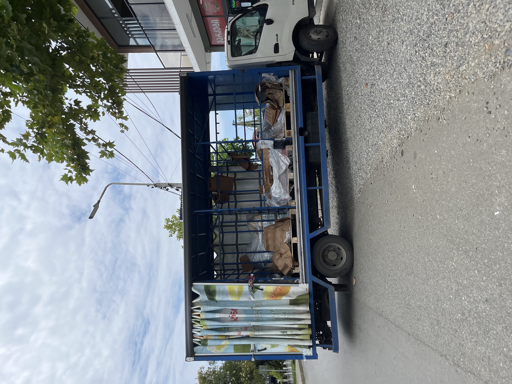 In Ureki, Georgia, a blue truck with an open cargo bed is parked on a grey asphalt road adjacent to a gravel shoulder. The truck's bed is filled with various items, including pallets stacked with goods wrapped in plastic and brown paper, as well as large white fabric bags. A blue metal cage structure is visible inside the truck bed. The truck has a white cab. To the right of the truck, a modern building with glass windows and a balcony is situated. A street lamp is positioned above and behind the truck. Trees with green leaves are visible in the background. The sky is overcast with clouds.