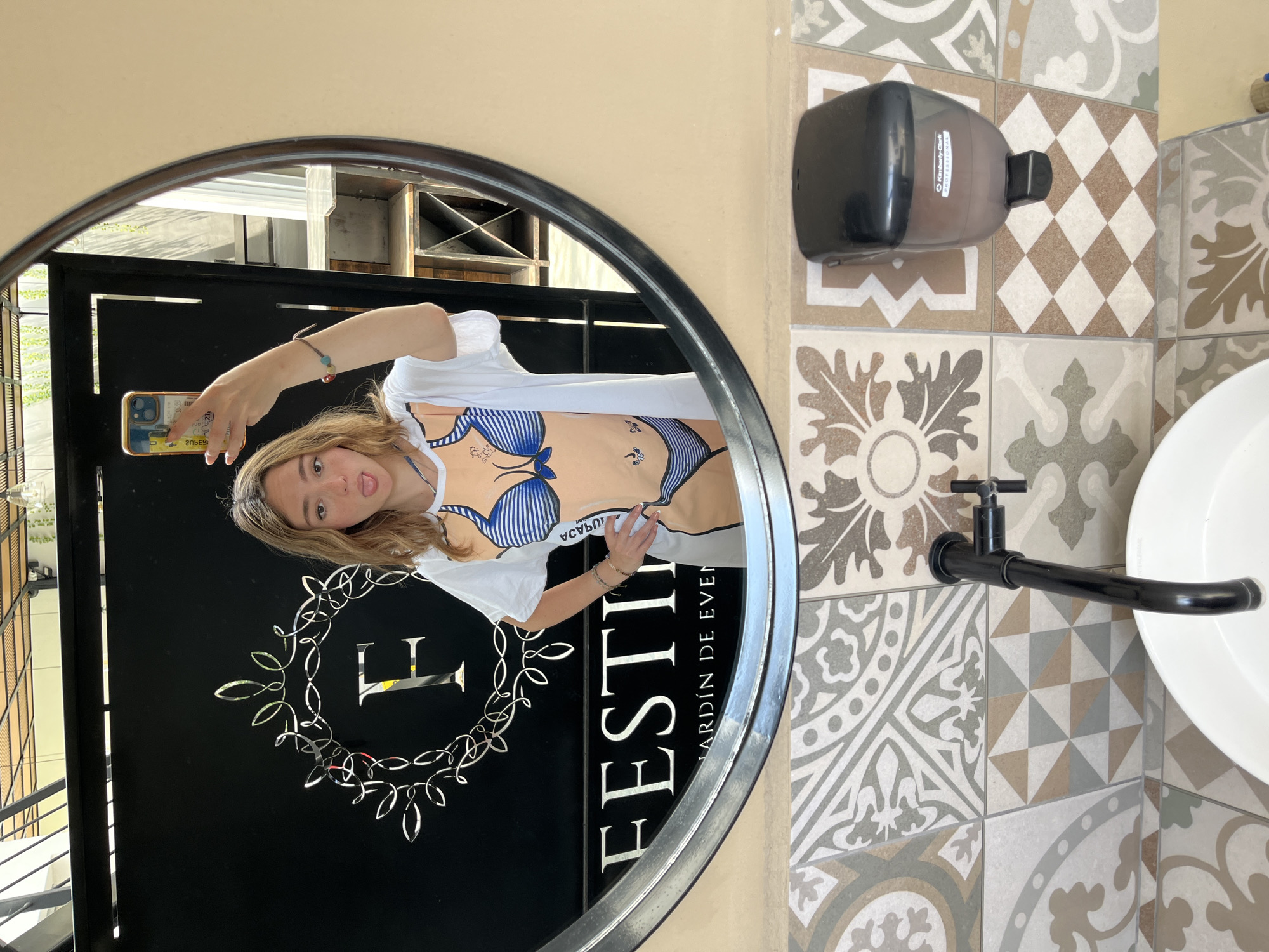 A blonde female takes a selfie in a circular mirror. She is wearing a white t-shirt over a top with a striped bikini print. The mirror reflects a black sign with the word "FESTIN" and "JARDIN DE EVENTOS" and an iron gate.
To the right of the mirror is a wall covered in decorative tiles. A black soap dispenser is mounted on the wall, and a black faucet and white sink are below.
The location is likely in San Bernardino Tlaxcalancingo, Mexico, based on context.