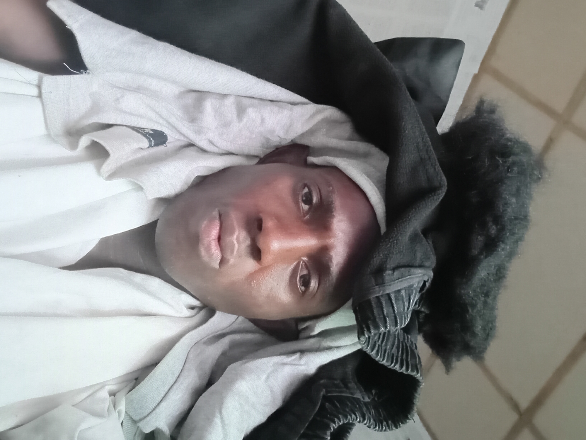 An overhead shot captures a dark-skinned individual lying among piles of clothing in a room. The person's face is the focal point, with eyes open and looking towards the camera. They are partially covered by several layers of clothes, including a white shirt, a grey garment, and a black item with what appears to be either a hood or a textured surface. The background is minimal, showing a light-colored wall with subtle vertical lines. There is no explicit indication of the location being Akwete, Nigeria in the image.