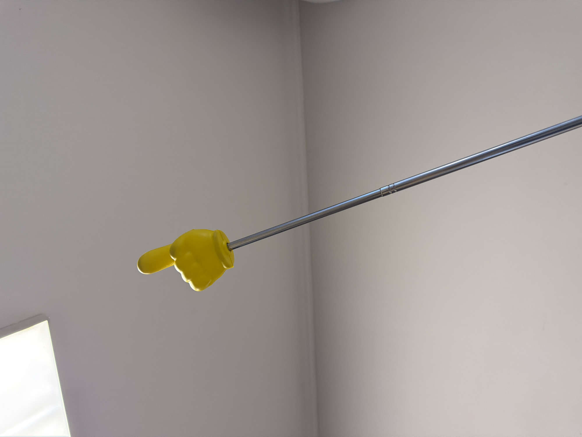 A yellow foam finger on a metal pole is pointed upwards against a light-colored wall and ceiling. A fluorescent light fixture is visible in the upper left corner. The scene is indoors and appears to be a room with painted walls and a dropped ceiling. No specific location in Yerevan, Armenia is identifiable.