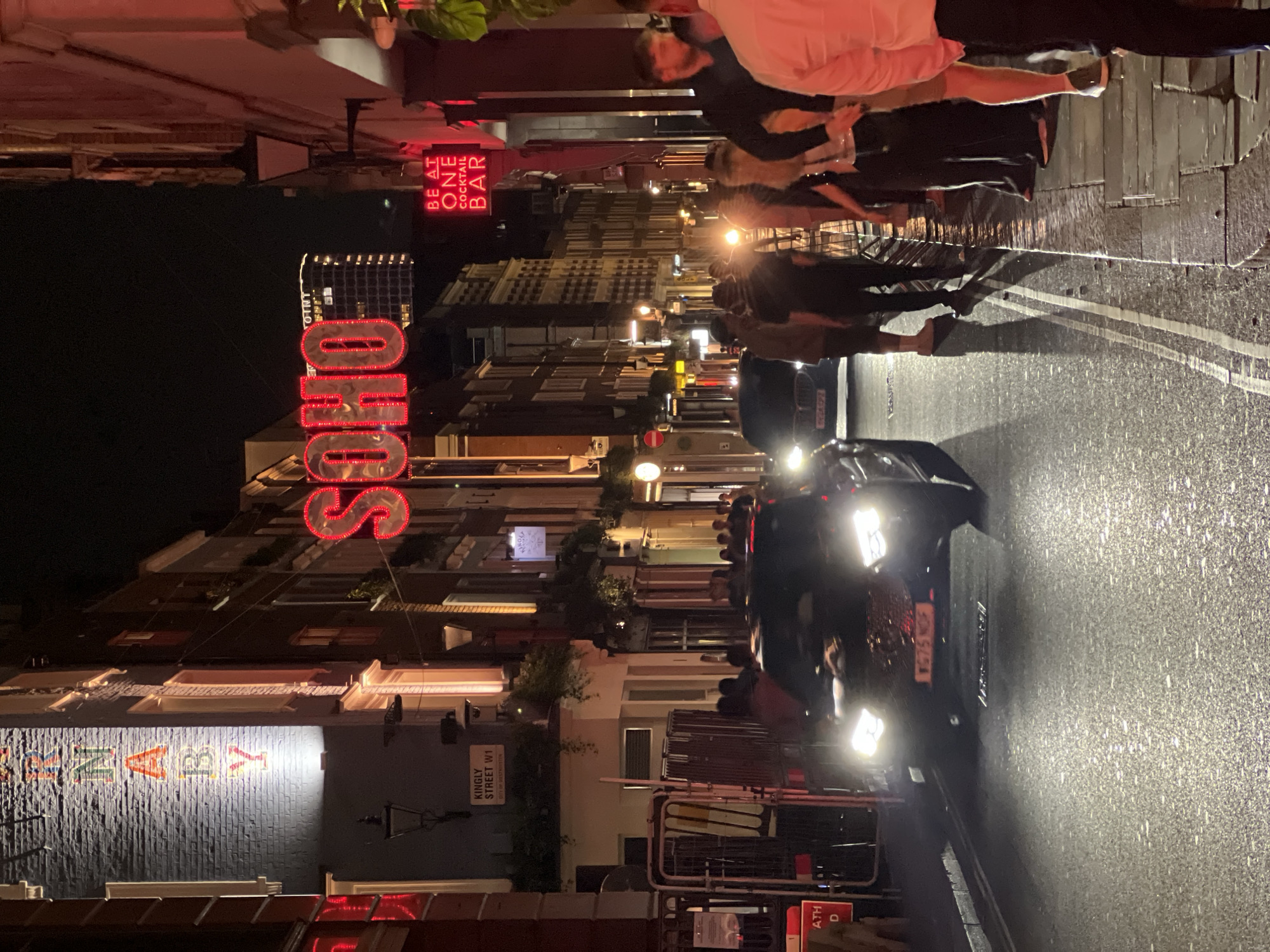 The image is a street scene taken at night in Soho, London. A dark-colored car with illuminated headlights and the partial license plate "VG75 NOF" is parked in the middle of the street. The street surface appears wet and reflects light. On the left side of the street, there is a building with a sign that reads "ZENAZY" in colorful block letters and above it, a large, illuminated "SOHO" sign. A street sign indicates "KINGLY STREET W1" on the same side. Above the Soho sign are multiple buildings and lights with people standing in front. There is another illuminated sign that reads "BE AT ONE COCKTAIL BAR" further up the street. People are seen standing and walking on the sidewalk on the right side of the street. The scene is generally illuminated by streetlights, building lights, and vehicle lights.