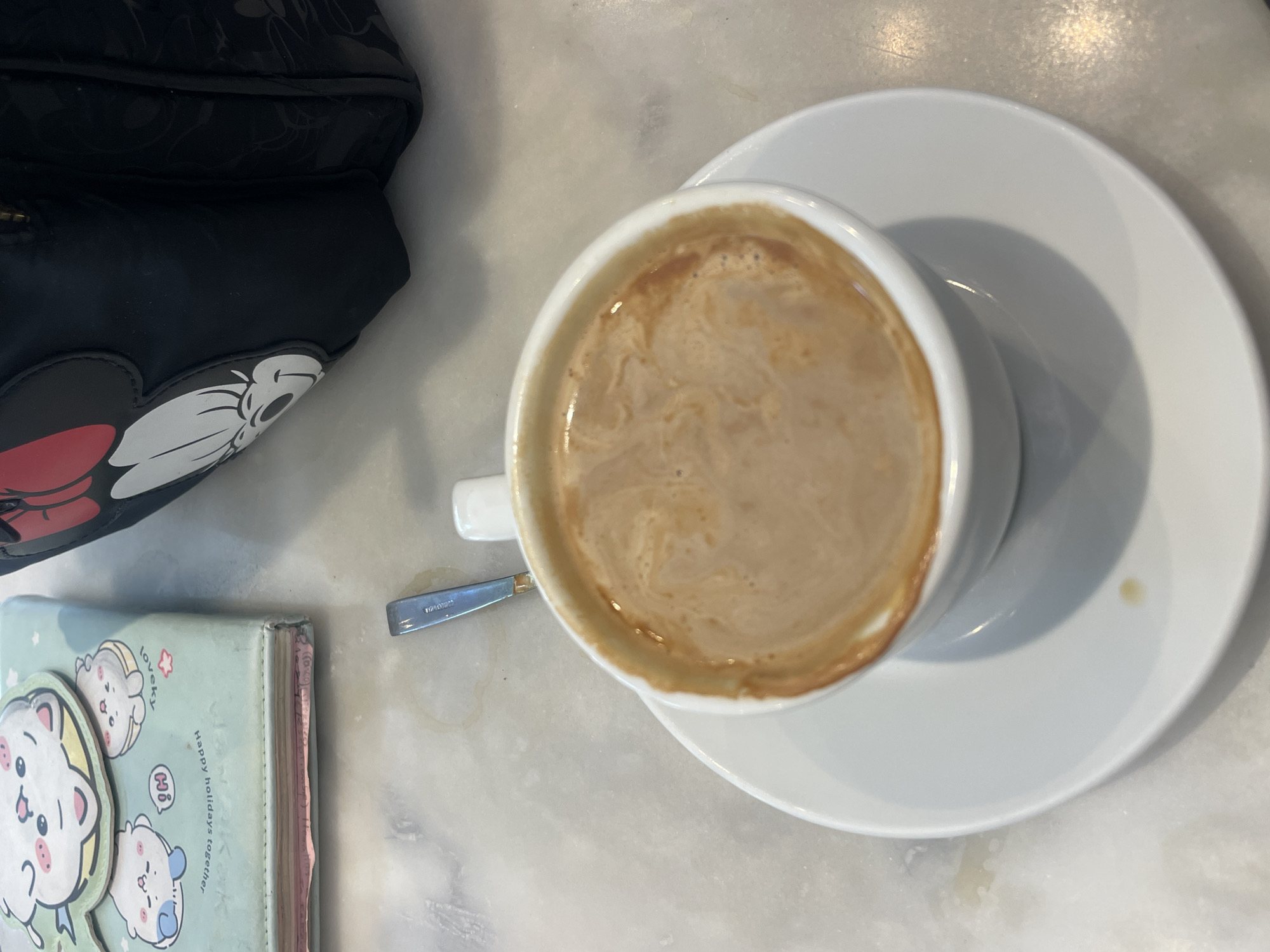 A close-up, overhead shot captures a cup of coffee with cream and foam resting on a white saucer on a marble-like tabletop. A silver spoon is partially visible to the left of the cup, resting on the table. In the background, a portion of a light blue notebook with cartoon animal illustrations and text is visible on the left, and a black bag with a Mickey Mouse design is on the right. The scene appears to be indoors, possibly in a cafe or restaurant in Barcelona, Spain, given the style of the items and the overall ambiance.