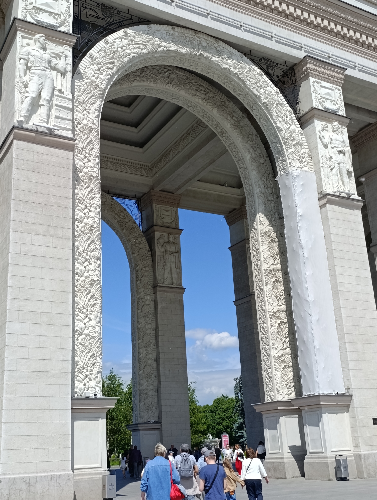 This vertical, low-angle photograph captures the monumental main entrance archway of the VDNKh (All-Russian Exhibition Center) in Moscow, Russia, on a bright, sunny day. The arch, constructed from light-colored stone or concrete, features ornate high-relief carvings characteristic of Soviet-era architecture.
The left pillar displays a relief of a male figure, possibly a worker, holding a tool or an agricultural implement. The arch itself is intricately decorated with abundant agricultural motifs, including sheaves of wheat, grapes, and various fruits, symbolizing prosperity and harvest. A stylized hammer and sickle emblem is subtly integrated into the decorative metalwork above the arch, consistent with the site's historical context.
On the right pillar, another relief figure, possibly a bare-chested male, is visible, also adorned with similar agricultural themes. A significant portion of the right arch support and pillar is covered in white protective or maintenance material, indicating ongoing restoration or upkeep.
Below the grand structure, several people, appearing to be visitors or tourists, are seen walking through the archway into the exhibition grounds. They are dressed in casual attire. The clear blue sky with scattered white clouds is visible through the arches, and green trees in the background suggest a vast park-like setting beyond the entrance. A grey trash bin with the visible text "ВДНХ" confirms the specific location. The strong sunlight casts clear shadows, highlighting the architectural details and suggesting midday.
