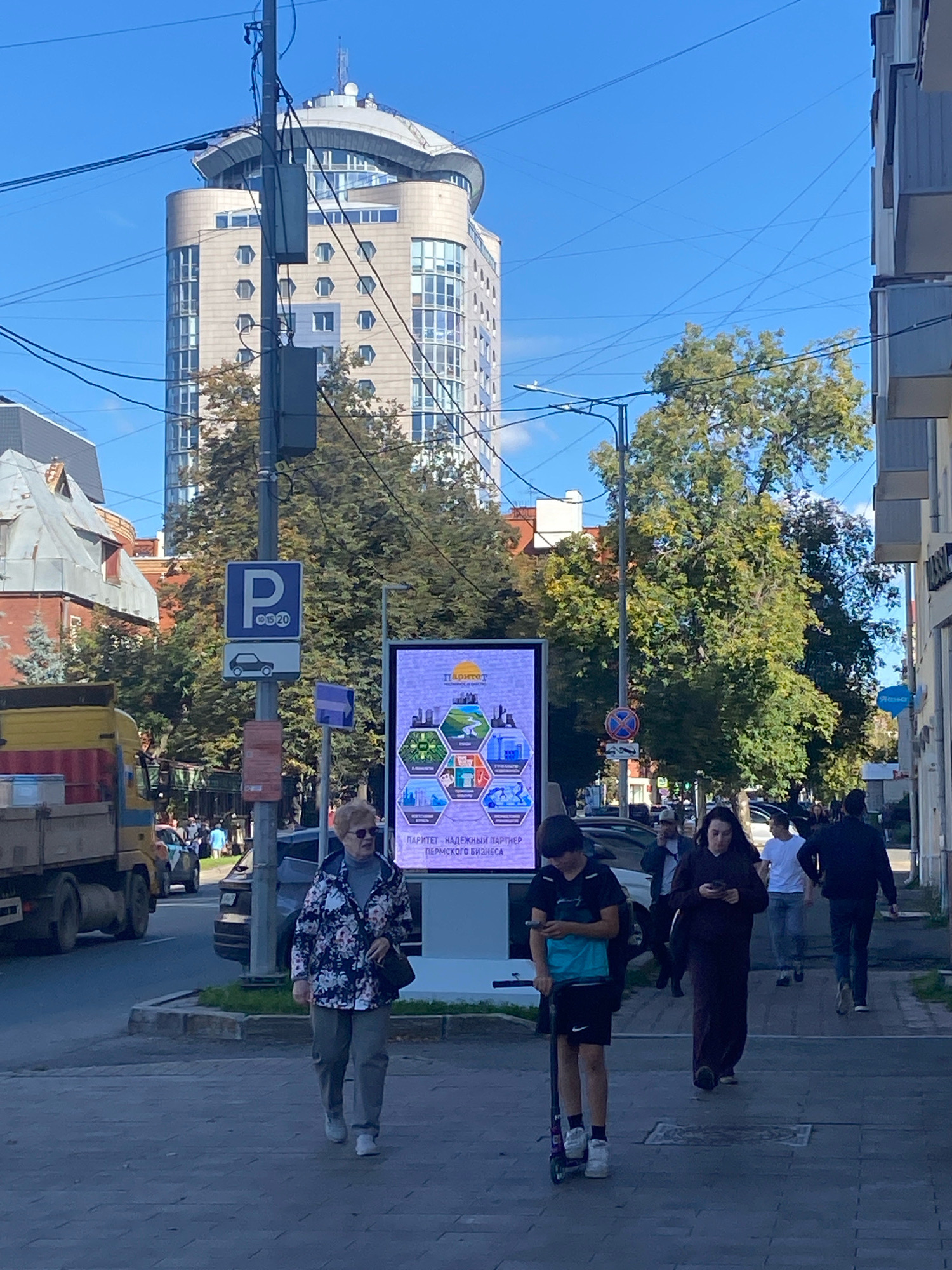 In Perm, Russia, on a sunny day, a sidewalk scene unfolds with a tall, modern building dominating the background. The building features a distinctive curved roof and many windows. A digital billboard displays a hexagonal infographic with icons representing industry, nature, and people, alongside Russian text that reads "ПАРТНЕР - НАДЕЖНЫЙ ПАРТНЕР ПЕРМСКОГО БИЗНЕСА" (Partner - Reliable Partner of Perm Business). A street sign indicates parking regulations with a 'P' symbol and numbers "10 5 20".
Several individuals are present. An elderly woman wearing a floral jacket and grey pants walks on the sidewalk, holding a dark purse. A young boy is on a scooter, looking at his phone. A woman in a dark robe walks while also looking at her phone. A man in a black jacket and jeans walks away from the camera. Other people are visible in the background, some walking and others near vehicles. A yellow truck is parked on the side of the street. Trees with green foliage line the street. The sky is clear blue.