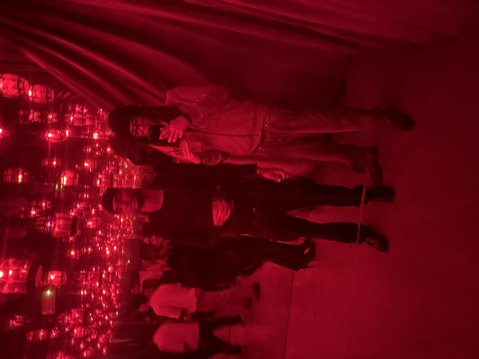 The image depicts an indoor immersive art installation or themed room, characterized by intense red lighting, located in Milan, Italy. The ceiling and walls are extensively mirrored, creating an infinite reflection of numerous glowing red lantern-like fixtures suspended throughout the space. A draped red fabric or curtain is visible along the right wall.
In the foreground, two male individuals are positioned, facing forward. The male on the left, with short dark hair, wears a dark long-sleeved top, dark trousers with a light-colored horizontal stripe near the ankles, and dark shoes. He holds a dark object, possibly a smartphone or drink, in his left hand at waist level. The male on the right wears a dark beanie, eyeglasses, a light-colored (appearing grey/beige under red light) zippered jacket, matching trousers, and dark shoes. He is actively taking a selfie using a smartphone, held with both hands in front of him, capturing himself and the adjacent male. In the blurred background, several other indistinct figures are visible, some standing and others in motion, consistent with a public exhibition space.