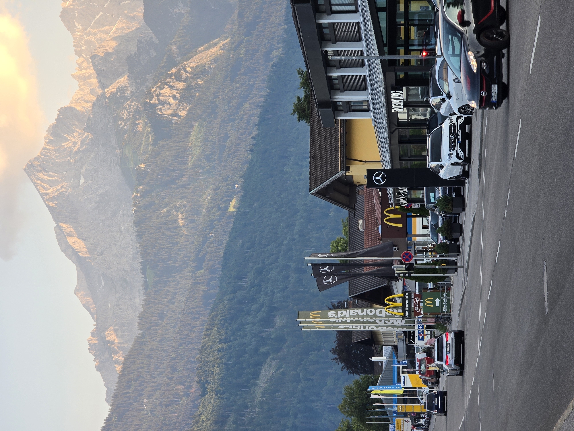 The image depicts a street scene in Garmisch-Partenkirchen, Germany, with a prominent mountain range in the background. The foreground shows a road with several cars, including a silver Audi in the left lane and a black car with red accents in the right lane. On the right side of the street, a Mercedes-Benz dealership is visible, with several cars on display, including a white Mercedes-Benz SUV. A McDonald's restaurant with its iconic golden arches is also present, along with tall yellow and black flags. The buildings in the scene have a modern architectural style. The mountain range in the background is steep and rugged, with forested slopes and snow-capped peaks, illuminated by the warm light of what appears to be either sunrise or sunset. The sky above the mountains is a pale blue with hints of clouds. The overall impression is a blend of urban infrastructure and the natural grandeur of the Bavarian Alps.
