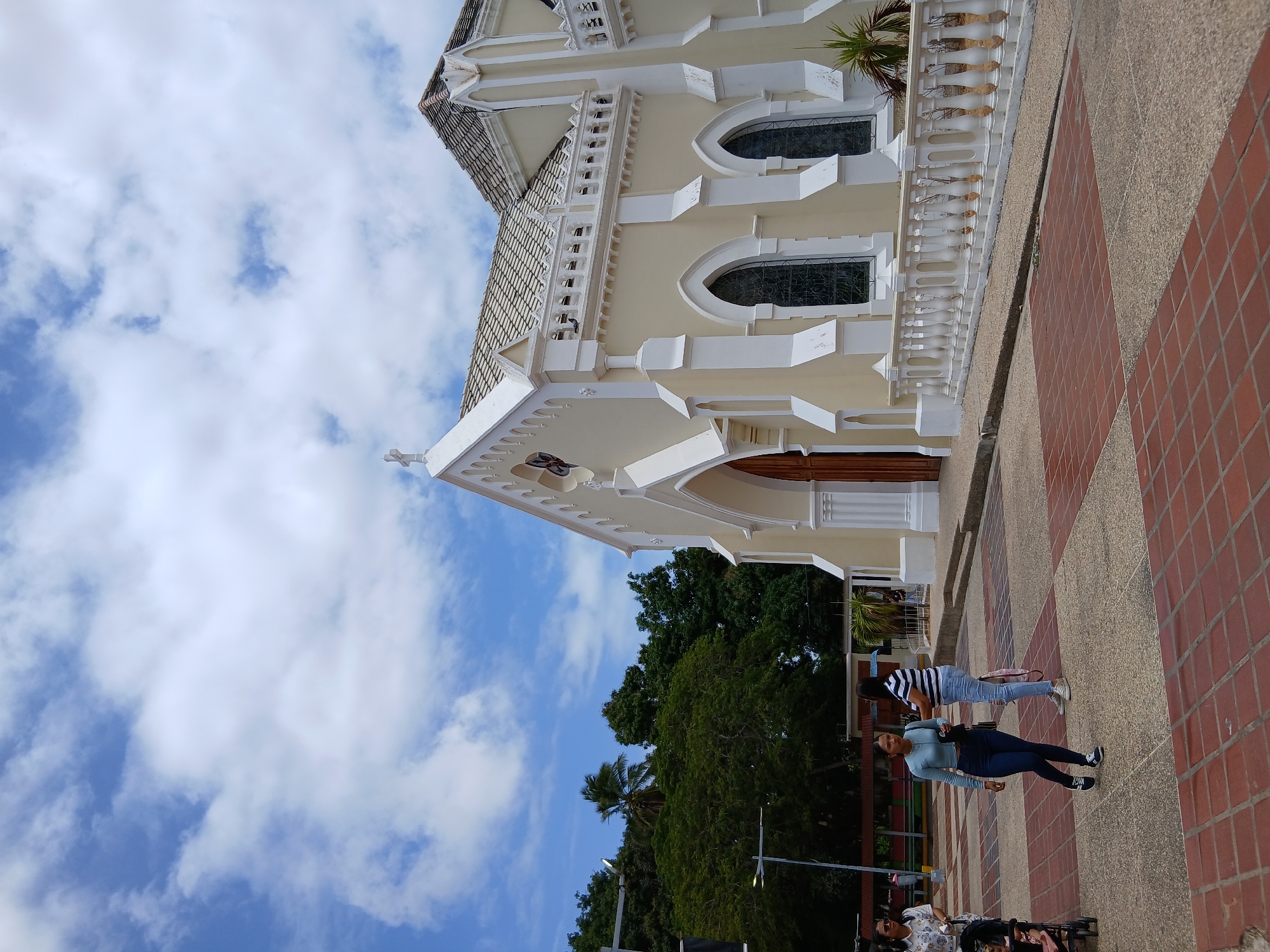 The image shows the Iglesia Inmaculada Concepción, a beige church with white trim, located in Altagracia, Venezuela. The church has arched windows and a tiled roof, and a cross on its front facade. Two women are walking on the tiled and concrete sidewalk adjacent to the church. In the background, there are trees and a blue sky with clouds. A group of people, including someone in a wheelchair, are partially visible in the lower portion of the image.