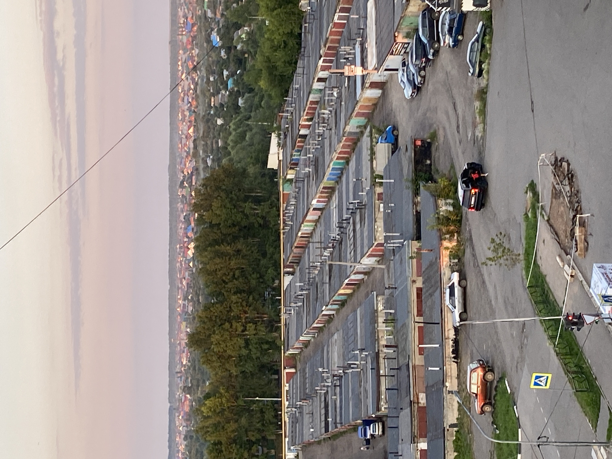 The image depicts a panoramic view of a suburban area with a dense collection of garages in the foreground, stretching towards a distant cityscape and rolling hills under a pale pink sky. The garages, arranged in long rows, have a uniform design with predominantly grey roofs and various colored doors, including red, white, and green. Several cars are parked in the lanes between the garages.
In the mid-ground, the landscape transitions to a neighborhood of houses with red-tiled roofs, interspersed with green trees. This residential area appears to be part of the city of Neftekamsk, Russia, as indicated by the context. Beyond the houses, a line of trees forms a dark green band against the horizon.
In the foreground, a street intersection is visible with a traffic light, a yellow pedestrian crossing sign, and a white road marking. A red SUV and a silver sedan are parked on the side of the road near some grass. Another black SUV with its rear door open is parked in a lot, with a person in a red outfit near it. A blue truck is parked in the vicinity of the garages. The overall scene suggests a residential area with extensive garage facilities, likely a common feature in cities like Neftekamsk.