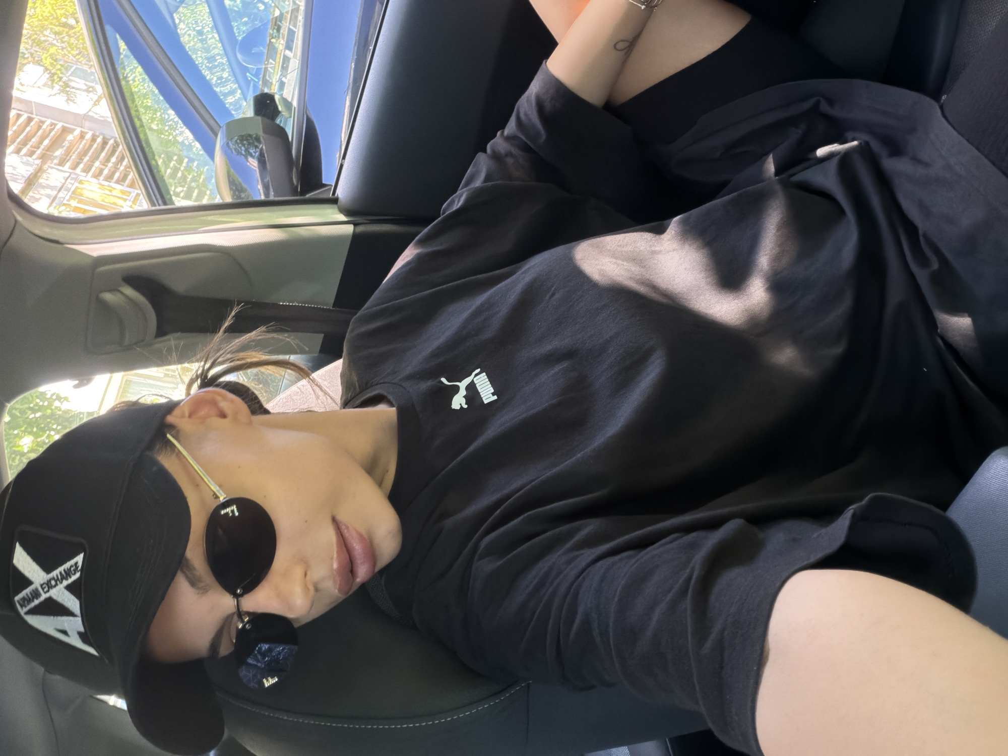 A woman is sitting in the passenger seat of a car, looking towards the right side of the frame. She is wearing a black baseball cap with a white logo that reads "AX ARMANI EXCHANGE", black oval sunglasses, and a black Puma t-shirt. Her hair is pulled back, and some strands are falling loosely. Her lips are slightly parted. She is wearing a silver bracelet on her left wrist and has a small tattoo on her forearm. The interior of the car is visible, including the black leather seat and part of the dashboard. Sunlight is streaming in, creating shadows on her clothing. Through the car window, a dark blue car and a building with windows are visible. The scene is outdoors, likely in a city setting like Burgas, Bulgaria, given the architectural style of the building and the presence of parked cars.