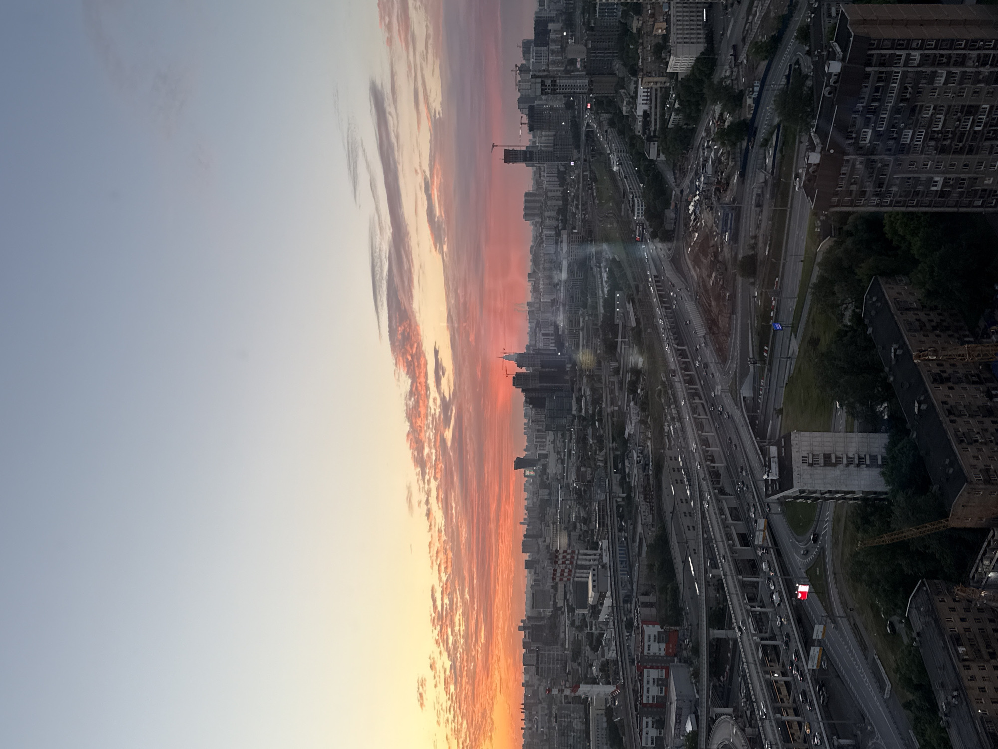 An aerial view captures a city skyline at sunset. The sky is a gradient of pale blue to warm orange and pink hues, with scattered clouds reflecting the light. Below, a dense urban landscape unfolds, characterized by numerous buildings of varying heights and architectural styles. A complex network of elevated highways and roads crisscrosses the scene, with cars visible as small lights on the thoroughfares. Some buildings appear to be under construction, indicated by cranes. The foreground shows residential buildings, some of which have visible windows and balconies. The overall impression is of a bustling metropolis at dusk, bathed in the dramatic colors of the setting sun.
