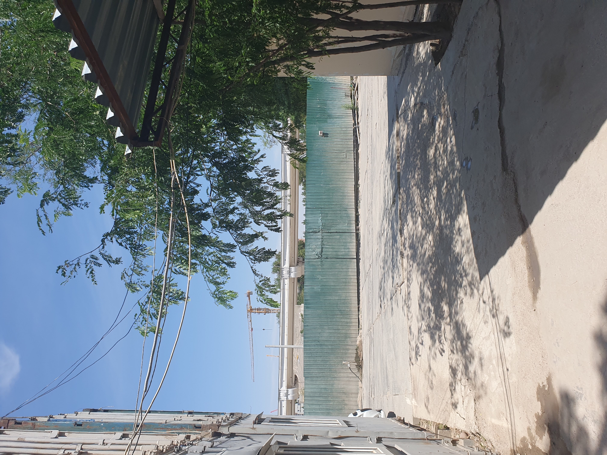 The image shows an outdoor scene with a light blue sky and some green trees. On the left, a light gray building, possibly a shipping container or modular structure, is visible. Electrical wires are strung across the scene, supported by poles and running past the building. A corrugated metal roof structure, possibly a temporary awning, is present on the right side of the image, casting shadows. In the middle ground, a long, light green corrugated metal fence runs horizontally across the frame, appearing to enclose an area. Beyond the fence, a section of elevated white piping or infrastructure is visible, with two construction cranes in the distance. The ground in the foreground is paved concrete with cracks and some puddles, and tree shadows are cast across it. The overall impression is that of an urban or industrial area under construction or in a state of transition. The scene is consistent with a location in Tashkent, Uzbekistan, given the architectural style and the presence of construction activities often seen in developing urban areas.