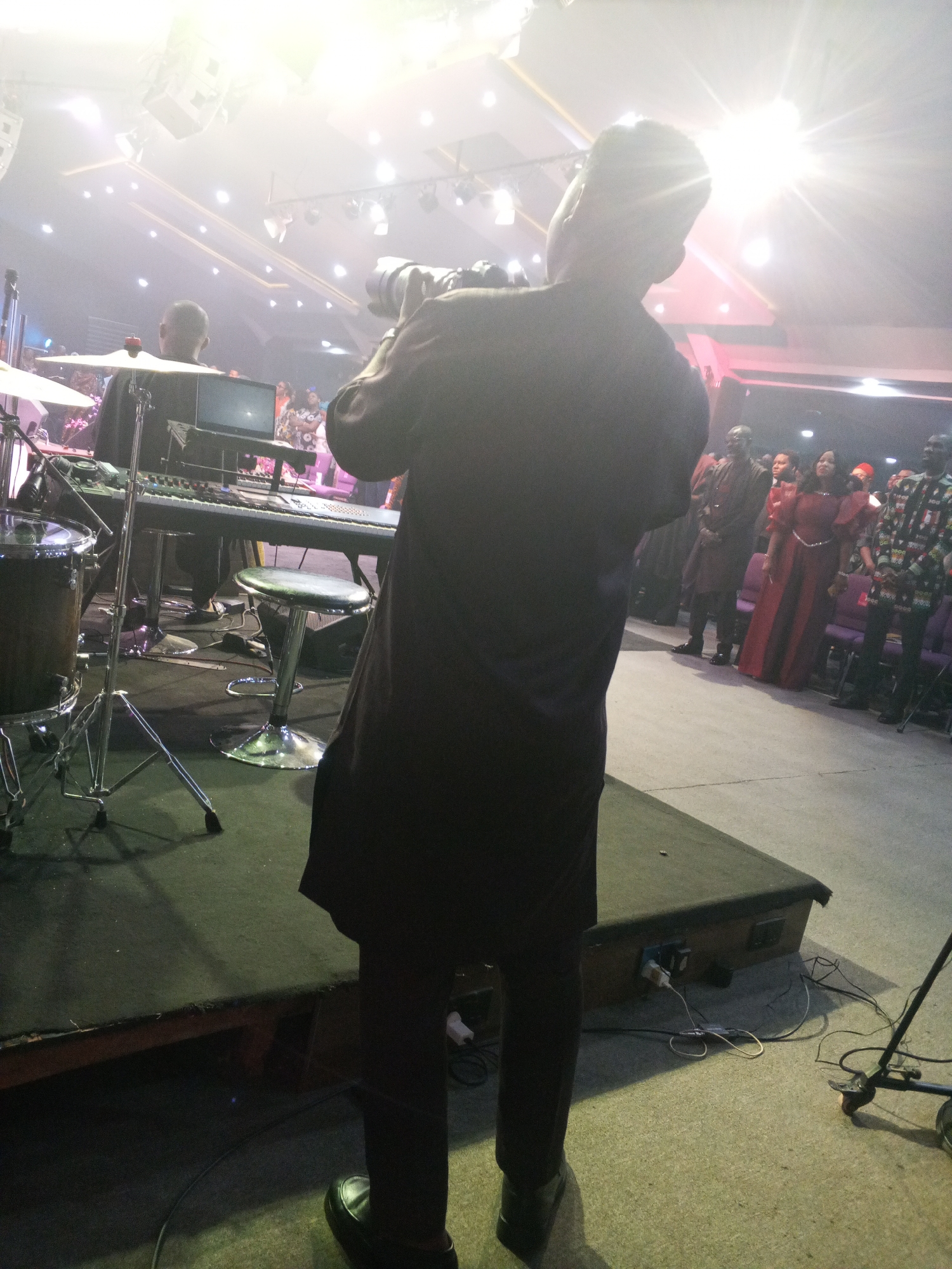 The image depicts an indoor event in Abuja, Nigeria, likely a performance or religious service. In the foreground, a person is seen from the back, holding a professional camera equipped with a large telephoto lens, aimed towards a stage. This individual is dressed in a dark, long-sleeved tunic over dark trousers and black shoes, standing on a light grey floor.
To the left, an elevated stage, covered in dark material, hosts musical equipment. A partial drum kit, including a floor tom and cymbal stand, is visible on the far left. In the center of the stage, a dual-keyboard setup rests on a stand, with a laptop and an audio mixer console positioned nearby. A person, likely a keyboardist, is seated on a high, chrome-legged stool behind the keyboards, seen from the back. Another similar stool is unoccupied to their right.
In the midground and background, an audience stands in rows, facing the stage. Several individuals are visible, including a woman in a long, bright red dress and a man in a patterned, multi-colored shirt. Purple chairs are partially visible behind the standing audience members.
The venue's ceiling is high, featuring numerous bright lights, some recessed and others appearing to be stage lights, which create significant lens flare and overexposure in the upper portions of the image. Speaker enclosures are mounted high on the walls. Near the stage edge in the immediate foreground, cables are visible, running over and under the stage, with power outlets attached to the stage structure. A wheeled microphone stand is positioned on the floor to the right of the foreground photographer.