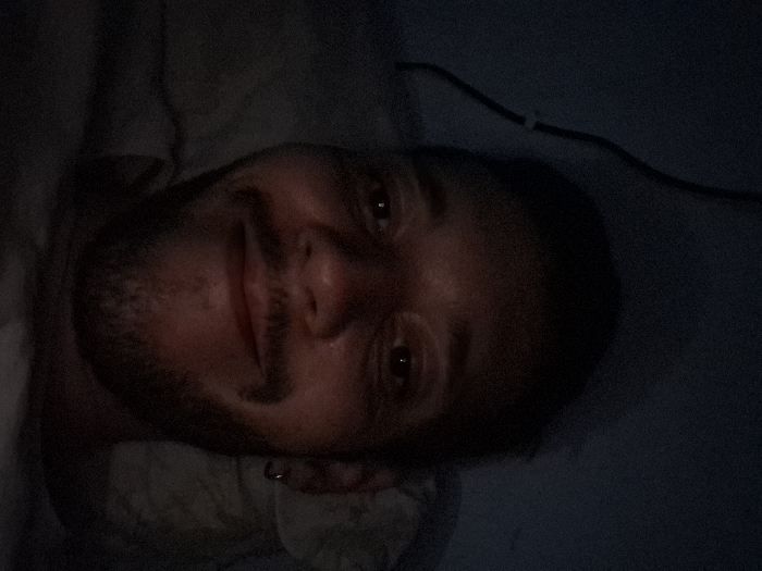 The image depicts a young man lying in bed, captured from the chest up. He is wearing a dark shirt. He is smiling and looking directly at the camera. The image appears to be taken in low-light conditions, as the environment is mostly dark. The man has short dark hair, a goatee, and is wearing a small circular earring in his right ear. The location of this image is in Angra dos Reis, Brazil, based on the provided coordinates.
