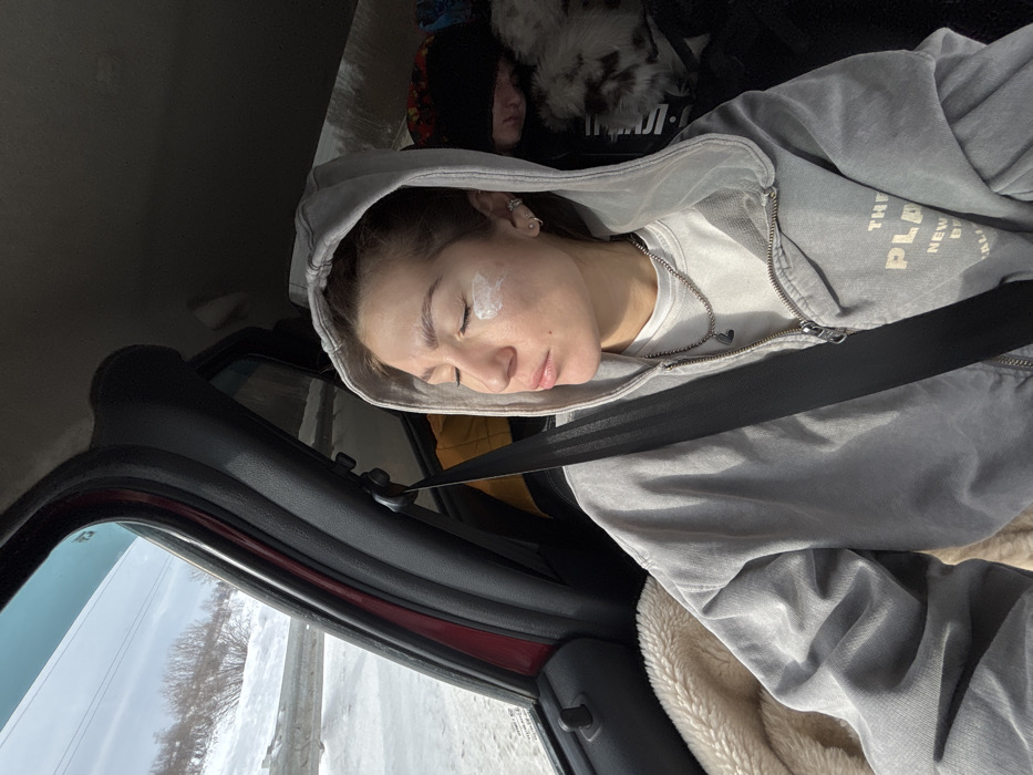 An image captured inside a vehicle depicts two individuals and a winter landscape. In the foreground, a young adult female is seated, secured by a black seatbelt across her chest. She wears a light grey hooded zip-up jacket over a white top and a silver chain necklace with a dark pendant. Her eyes are closed, and a visible dollop of white cream is present on her left cheekbone. Multiple small hoop earrings adorn her left ear. To her left, a cream-colored, fuzzy textile is visible. In the background, to the right of the primary subject, a second individual, possibly a child, is partially visible, appearing to be asleep. This individual wears a black beanie, and a white and brown patterned object, possibly a blanket or animal fur, rests near them. Behind this second individual, a silver, reflective material is present, likely a sunshade or insulation. Through the car window on the left, a snow-covered outdoor environment is visible, featuring bare trees or shrubs on a gentle slope, under a bright, clear sky. A metal guardrail is partially discernible along the roadside. The scene is located within the city of Ustinovo, Russia.