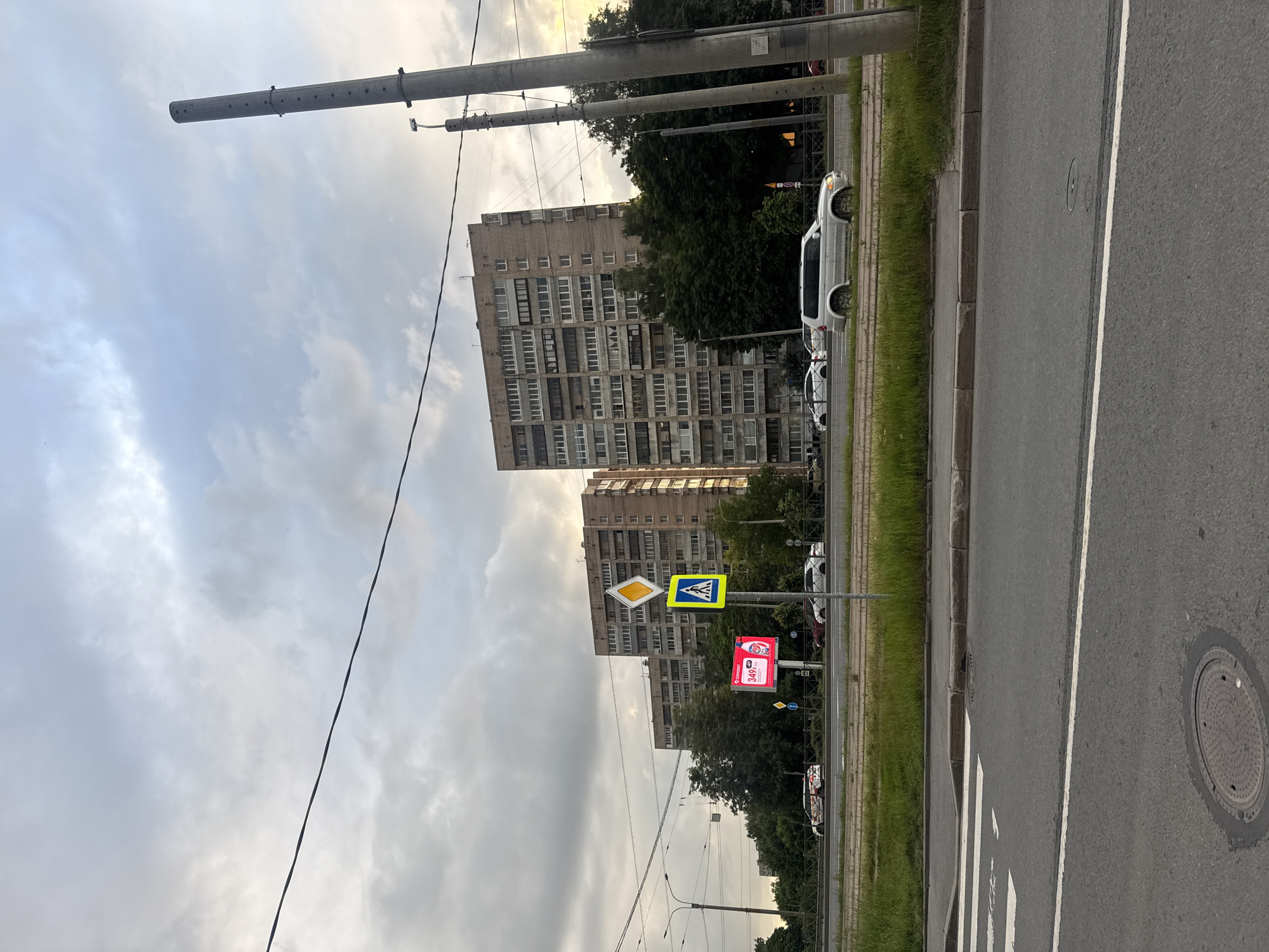 The image depicts an urban street scene in Kokkorevo, Russia, with a focus on a multi-lane road and residential buildings. The sky is overcast with clouds. On the left side of the road, a pedestrian crossing is marked by white lines. A large digital billboard displaying pink advertisements is visible in the middle ground. Adjacent to the billboard, traffic signs, including a pedestrian crossing sign and a diamond-shaped warning sign, are mounted on a pole. Several cars are driving on the road, including a silver SUV on the right. Behind the road, a line of trees separates the road from a row of tall, multi-story apartment buildings with numerous windows. Two large concrete utility poles stand on the right side of the image, one prominently in the foreground. Power lines are strung across the sky. The overall impression is of a typical day in a Russian city, with infrastructure and residential life coexisting.