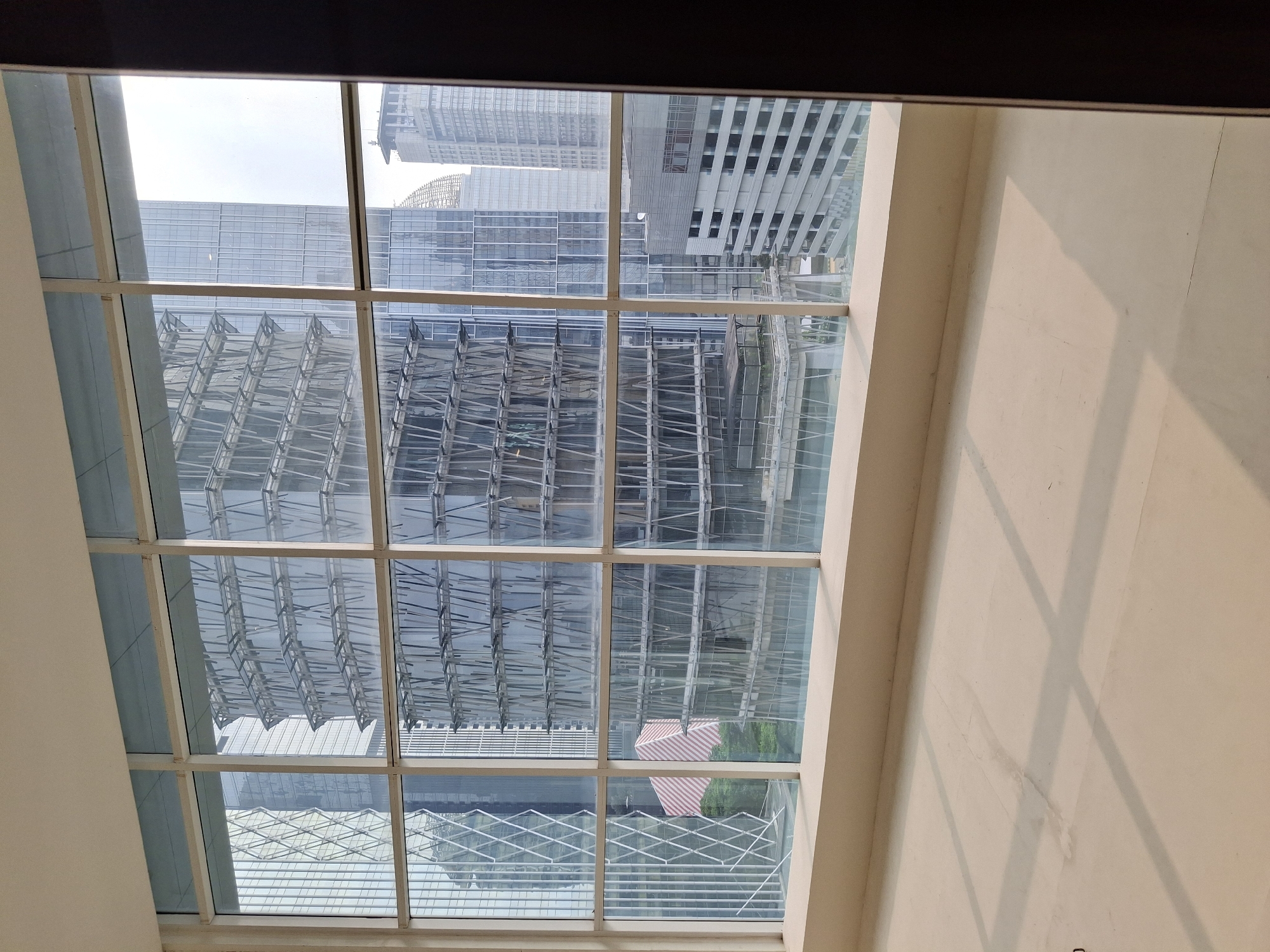 The image shows a view from inside an empty room, looking out through a large, multi-paned window at a cityscape. The window frames are white and divide the view into a grid.
Outside, several modern skyscrapers are visible. The central building is a tall, cylindrical structure with an intricate, crisscrossing facade made of glass and metal supports. To its left, another building has a geometric pattern of diamond shapes formed by its exterior structure. To the right of the central building, other skyscrapers of varying heights and architectural styles are present, some with glass facades and others with more opaque exteriors. One building in the distance has a distinctive domed or curved top.
In the foreground, inside the room, the floor is a light-colored, smooth surface, possibly concrete or a similar material. Sunlight casts distinct rectangular shadows across the floor, indicating the direction of the light source. The wall at the bottom of the window is a light beige color. The right edge of the image shows a dark brown vertical element, likely a door frame or a wall.
The scene appears to be from a high-rise building, offering a panoramic view of a densely developed urban area. Given the architectural style of the buildings and the overall context, this scene is likely located in a modern business district. Based on the provided information and visual cues, the location is identified as South Jakarta, Indonesia.