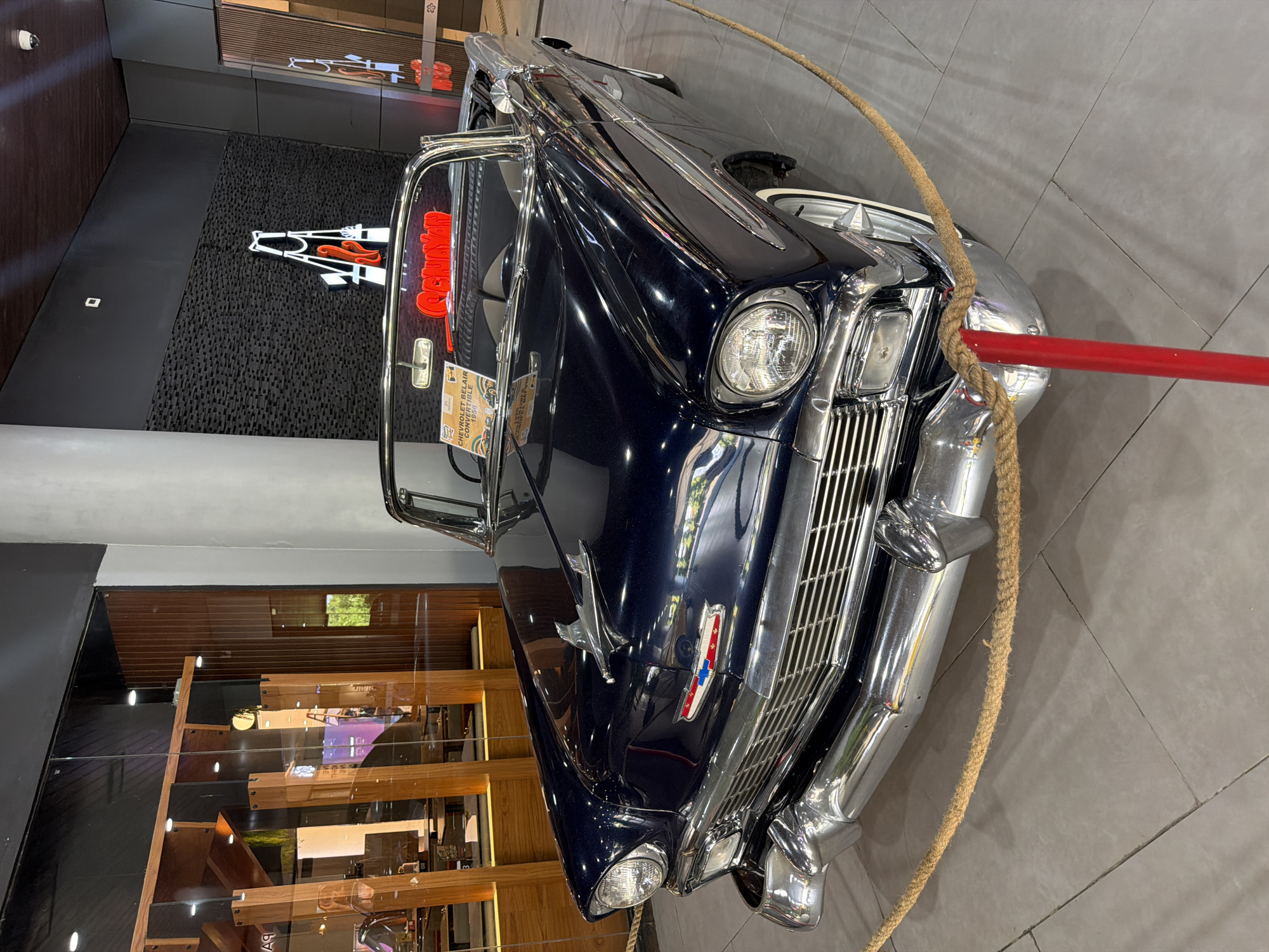 An antique dark blue Chevrolet Bel Air Convertible from 1956 is the central subject, positioned indoors on a grey tiled floor and cordoned off by a thick rope and a red barrier pole. The car is pristinely kept, showcasing its glossy dark paint, extensive chrome detailing on the grille, bumpers, headlight bezels, and side trim, along with white wall tires. An informational plaque clearly stating "CHEVROLET BEL AIR CONVERTIBLE 1956" is attached to the windshield wiper.
The setting appears to be an upscale showroom, museum, or a commercial space like a shopping mall, given the polished environment. Behind the car, a black, textured wall features a distinctive white and orange neon sign, which appears to spell out "GEHANI" or a similar word. On the left side of the frame, there's a modern wooden structure with glass panels, possibly a staircase or an elaborate display, which is also reflected in the car's highly polished surface. A security camera is visible on the upper left wall, indicating the valuable nature of the display. The overall scene suggests a curated exhibition in Kuta, Indonesia, with the classic car being the focal point.