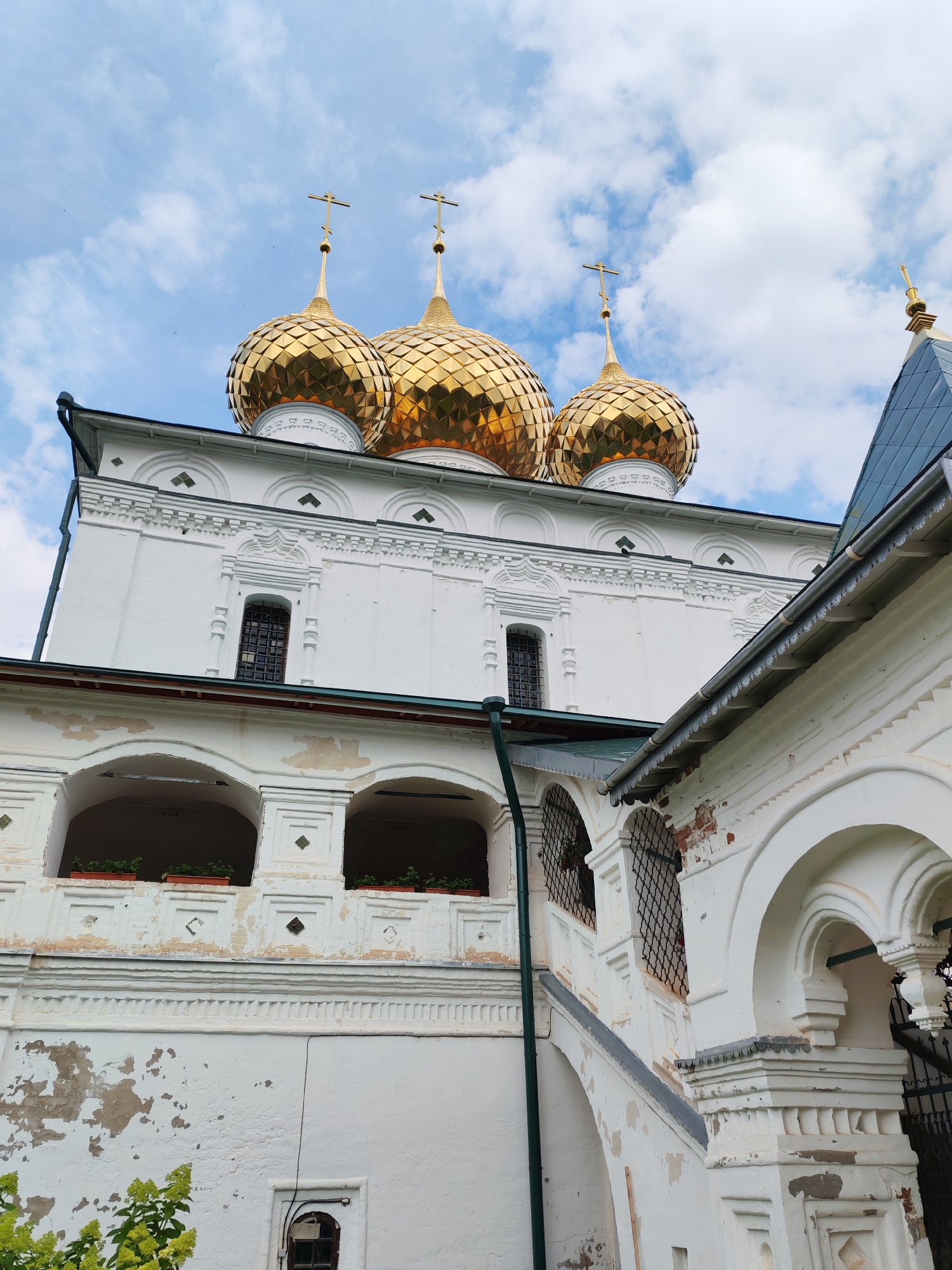 The image shows an exterior view of a white church with golden domes. Three prominent golden domes with crosses on top are visible against a blue sky with scattered clouds. The church has white walls with decorative architectural elements, including arched windows and cornices. Some parts of the building show signs of age and weathering, with peeling paint. In the foreground, there are white arched structures, possibly part of a cloister or walkway, with decorative grating and some greenery visible. The overall scene is one of religious architecture under a cloudy sky. The image is a photograph taken from a low angle, looking up at the church.