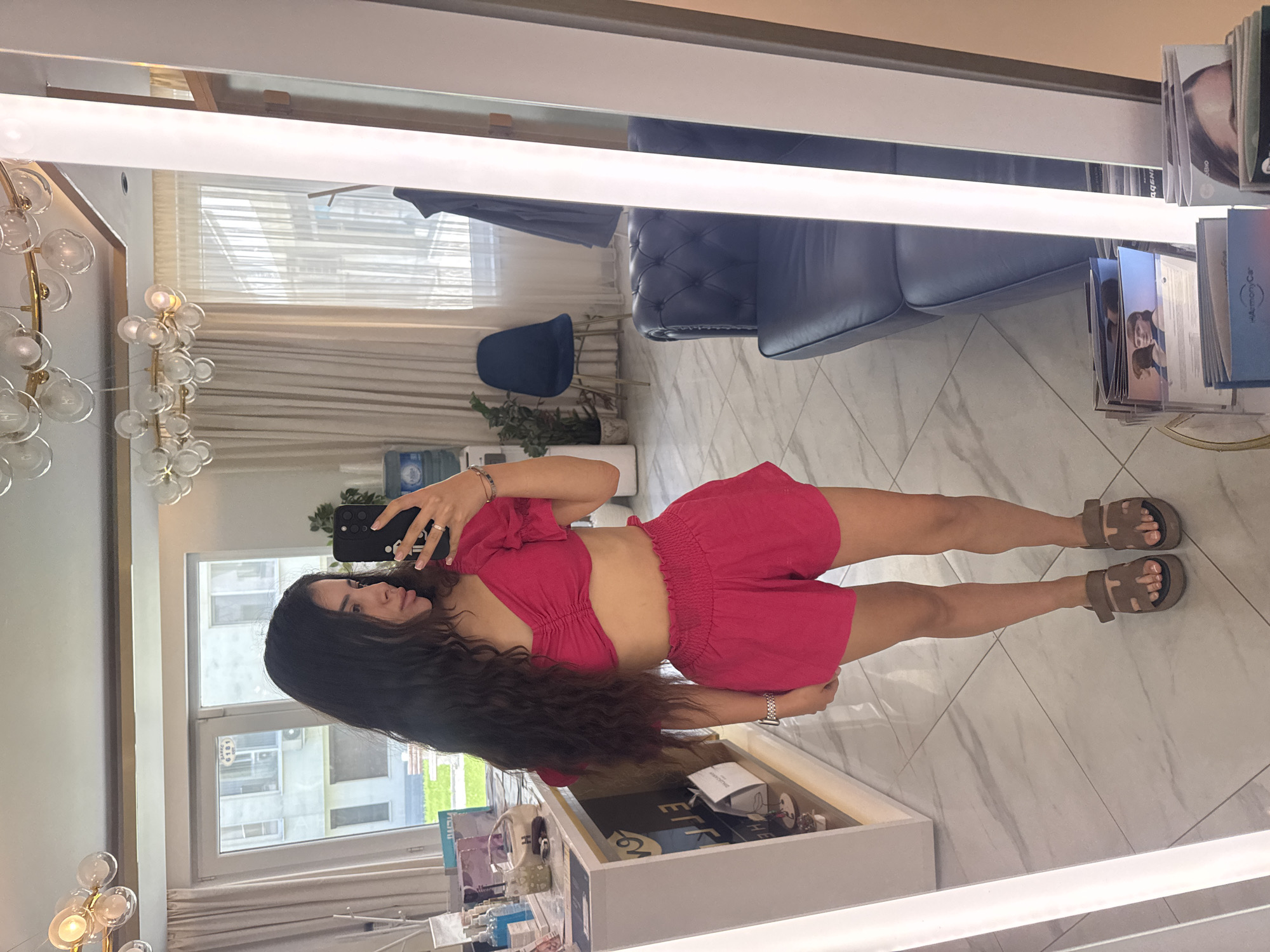 A young woman with long, dark, wavy hair is standing in a brightly lit room, reflected in a large mirror. She is wearing a pink, ruffled crop top and matching high-waisted shorts. Her right hand is holding a black smartphone, which she is using to take a selfie. She is also wearing brown platform sandals and a watch on her left wrist. The room appears to be a modern salon or spa, with a tiled floor, a leather sofa, a potted plant, and a decorative chandelier on the ceiling. In the background, through a window, a cityscape can be seen. The woman is positioned in the foreground of the mirror's reflection, occupying the central part of the image. The location is a beauty salon on the ground floor of a building on the main pedestrian street, Knyaz Alexander I, in the city of Plovdiv, Bulgaria.