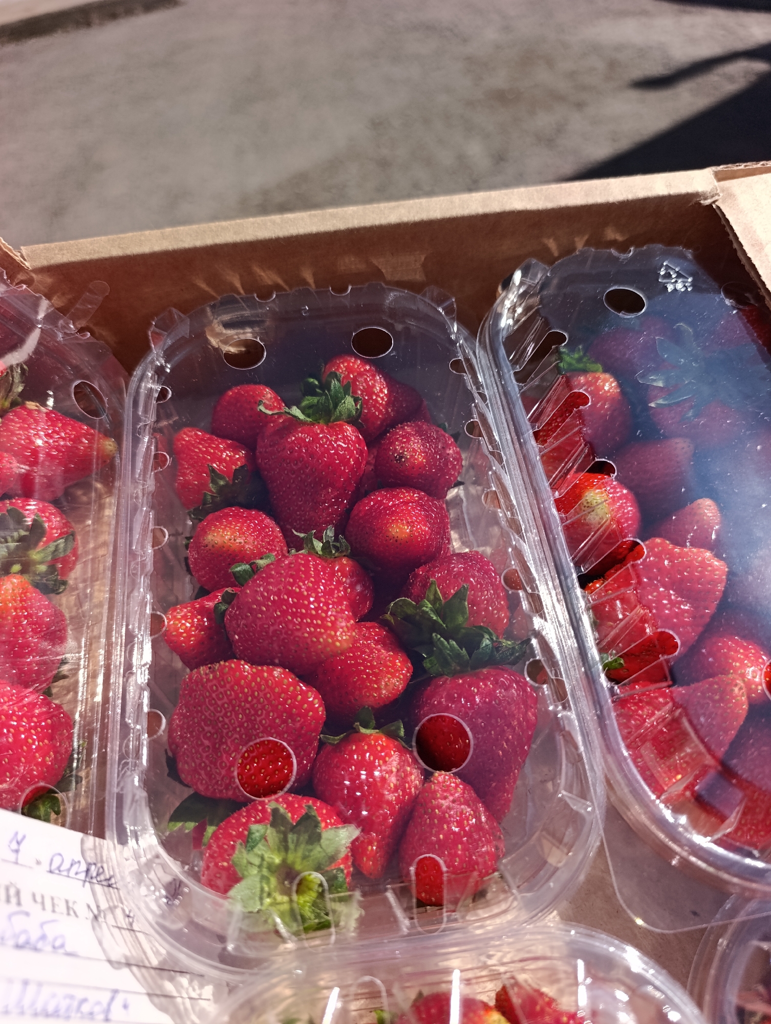 The image shows a cardboard box containing several clear plastic containers filled with fresh strawberries. The strawberries are bright red and have green stems. One container has a handwritten note attached to it. The environment appears to be a sunny outdoor setting. While the image suggests a market or sale of strawberries, I cannot confirm the location in Krasnoyarsk, Russia, based on the given information.