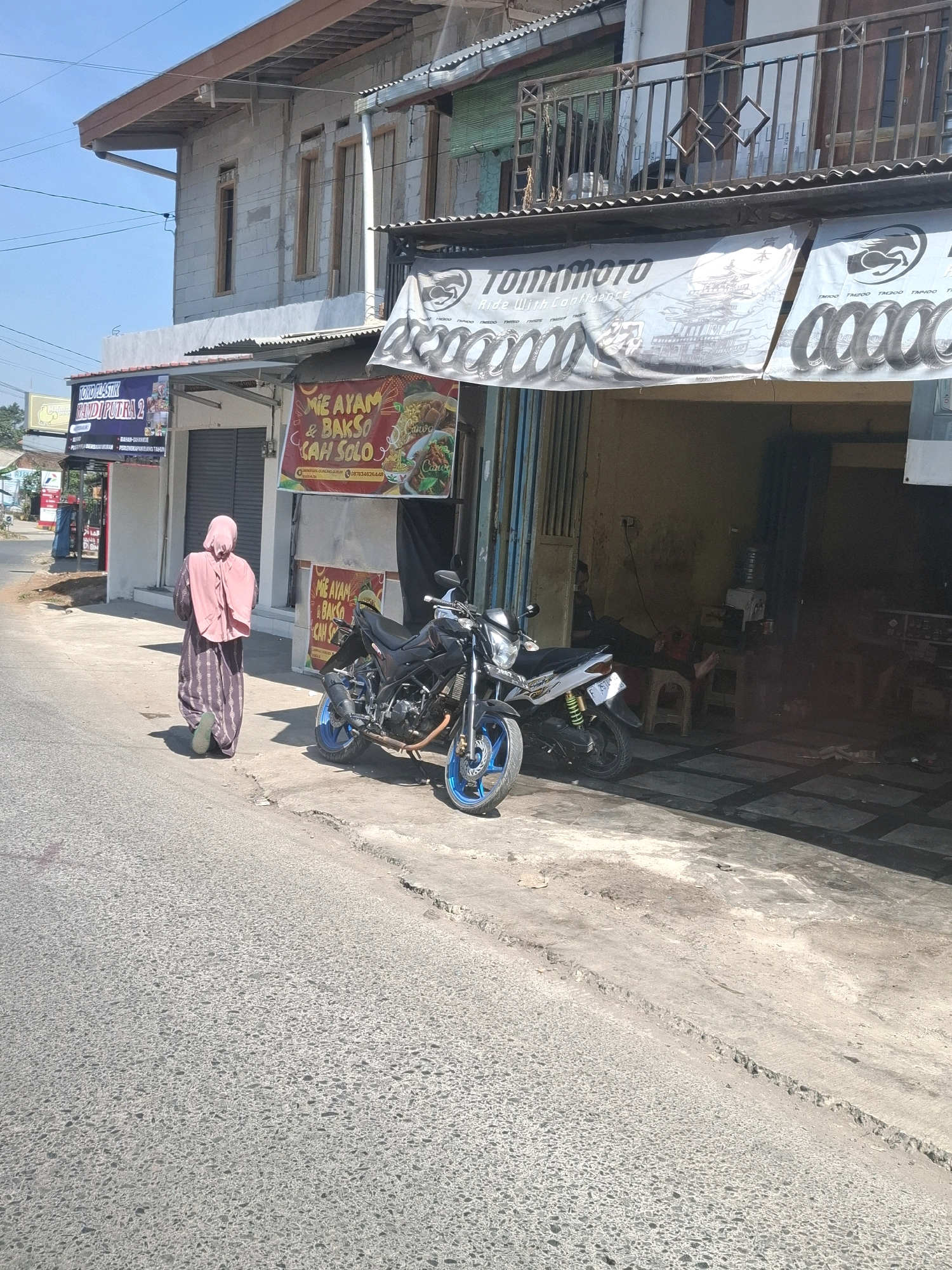 A street-level view shows a building with shops at the ground floor and a balcony on the second level, situated in Gunung Guruh, Indonesia. The building features a gray brick facade, with wooden window frames visible. A blue metal railing encloses the balcony. In front of the building, a banner with the text "TOMIMOTO RIDE WITH CONFIDENCE" and images of tires is displayed horizontally. Another banner advertises "MIE AYAM & BAKSO CAH SOLO". To the left, a sign for "TOKO PLASTIK HANDIPUTRA 2" is visible. Two motorcycles, one black and one white with blue accents, are parked in front of the shops. A person wearing a pink hijab and a long patterned dress is walking away from the viewer down the street. The street is paved with asphalt and concrete, with some cracks and uneven surfaces. The sky is clear and blue.