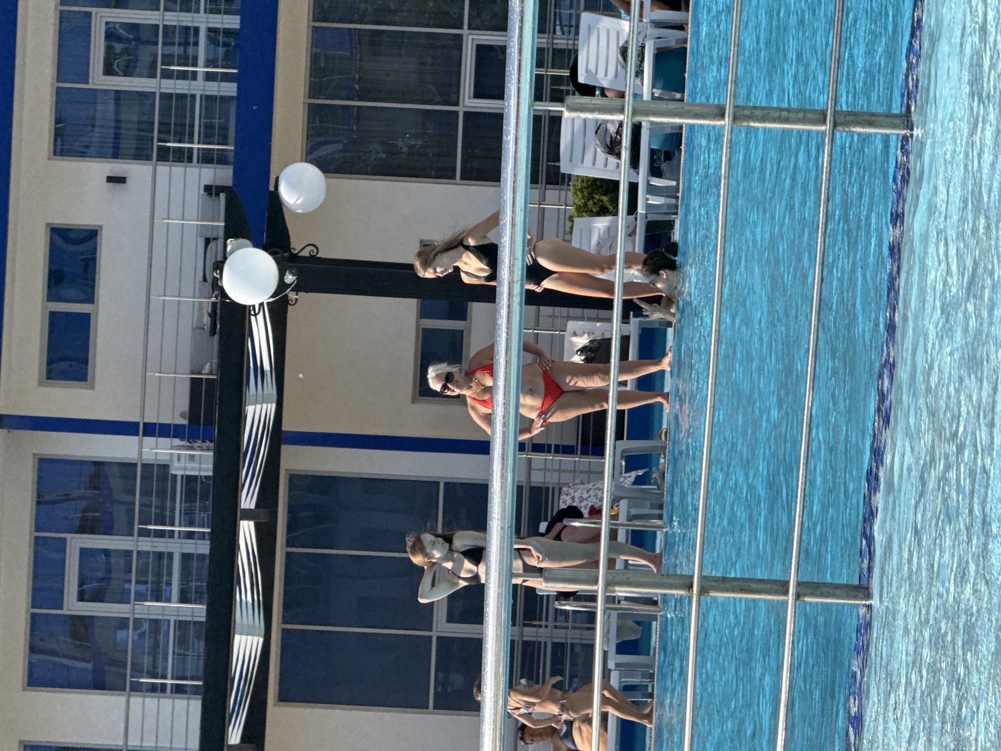 Three women are standing by a swimming pool. One woman in a black bikini has her hand on her head and is looking down. Another woman in a red bikini stands with her hands on her hips. A third woman in a black swimsuit stands looking to her right. The building behind them is white with blue trim and has balconies with glass doors. There are white lounge chairs and plants around the pool. The water is a bright blue color. The scene is a resort in Sevastopol, Ukraine.