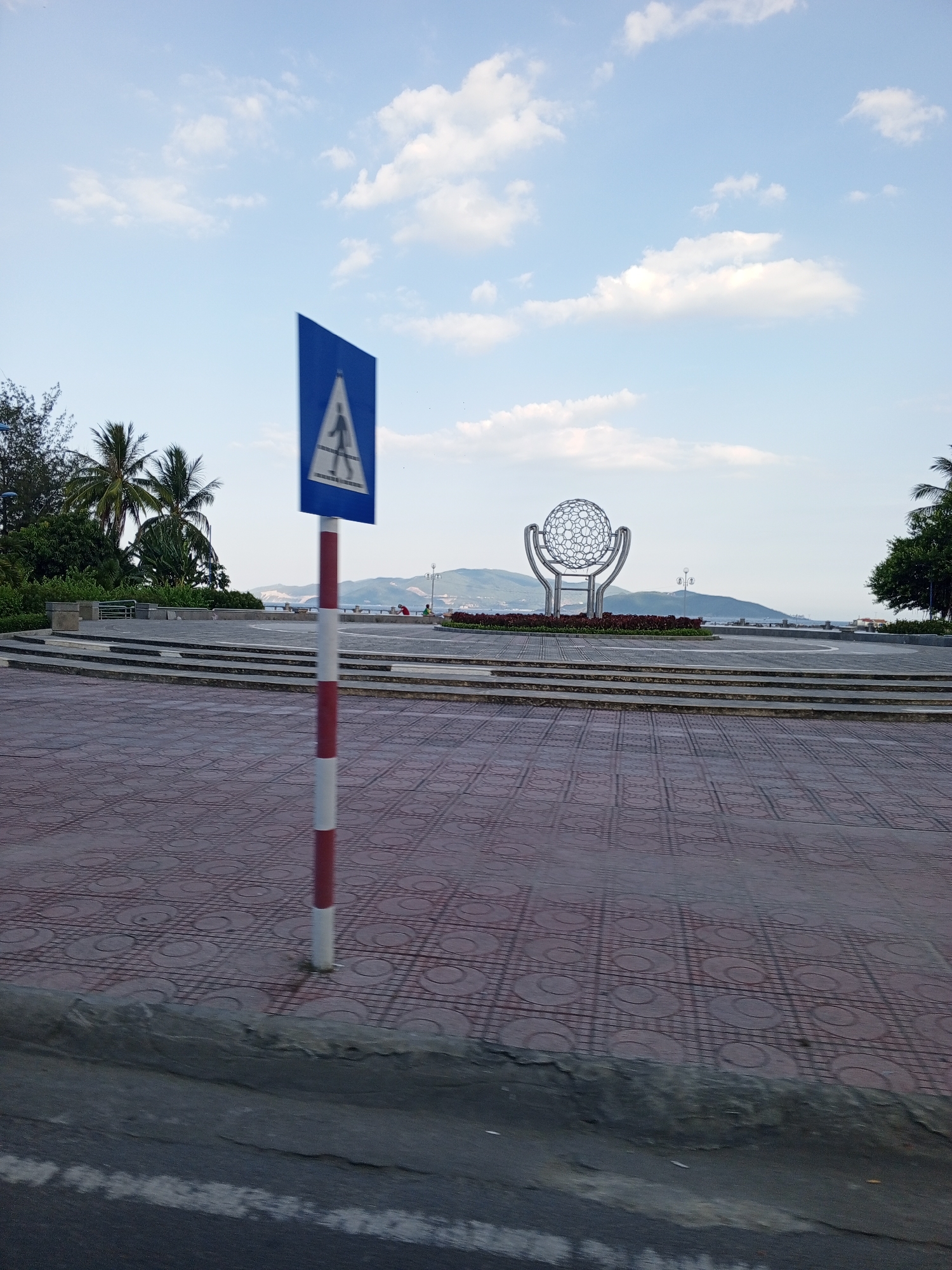 The image depicts an outdoor scene in Bac Nha Trang Ward, Vietnam. The foreground is dominated by a patterned red tile walkway and a section of asphalt road. A red and white striped pole stands on the walkway, topped with a blue triangular pedestrian crossing sign featuring a white pictogram of a person walking.
In the mid-ground, a circular metal sculpture, resembling a globe or sphere made of intricate latticework, is the focal point. It is surrounded by a flowerbed with dark red foliage. Behind the sculpture, there are tiered stone steps leading up to a paved area with some benches and lampposts. In the background, rolling hills and mountains are visible under a bright, partly cloudy sky. Some palm trees and other greenery suggest a tropical or subtropical environment. The overall setting appears to be a public square or park area near a waterfront, given the hint of water and distant structures across the bay.
The time of day appears to be late afternoon or early evening, indicated by the soft lighting and the angle of the sun. The weather seems pleasant, with scattered clouds in a clear blue sky. There are a few people visible in the mid-ground, including one person in red sitting on a bench and another person standing. Their presence suggests a calm, leisurely atmosphere. No visible text is present in the image.