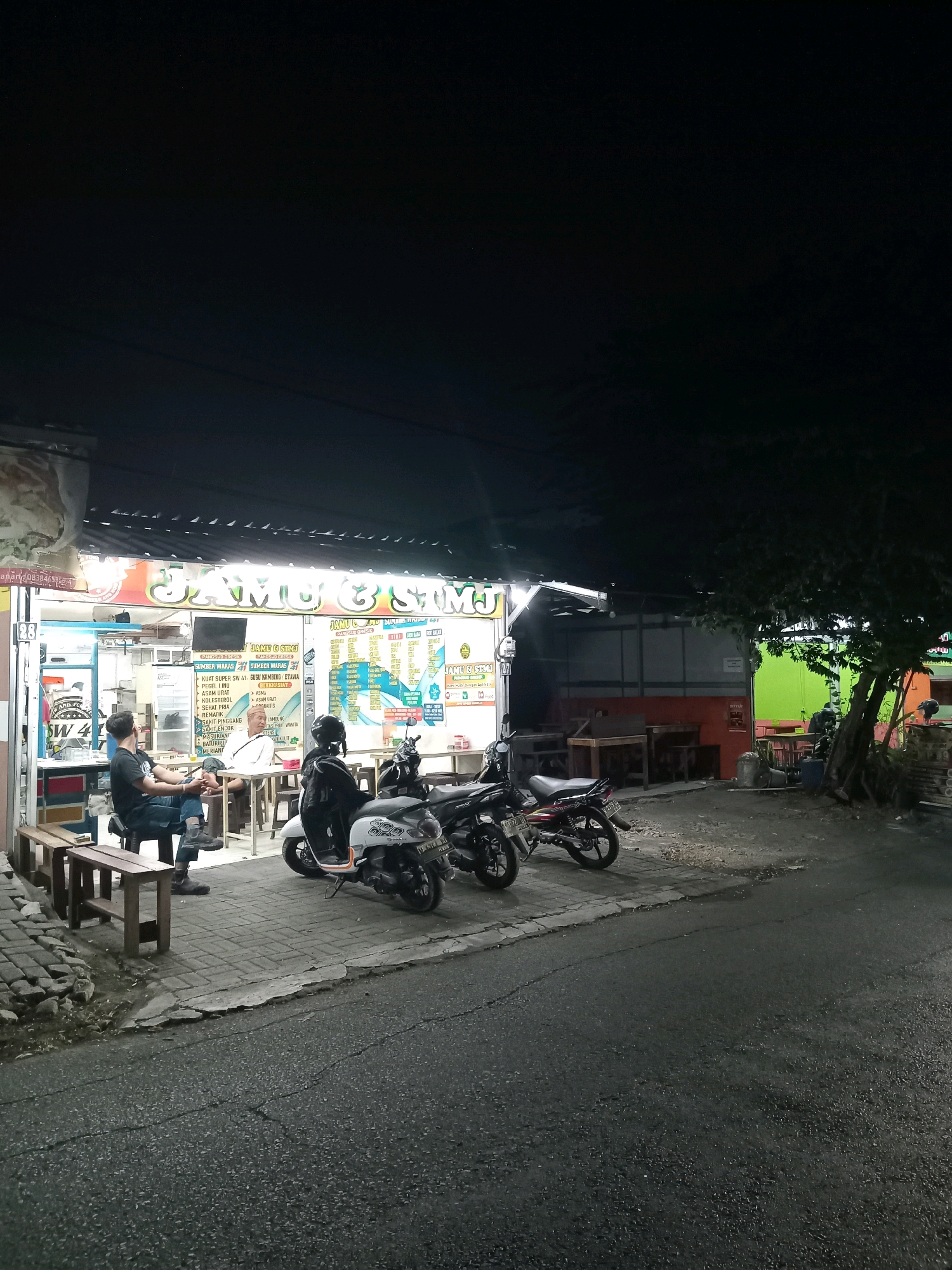 The image depicts a street-level view of a storefront at night in Randuagung, Indonesia. The storefront, illuminated by neon signs, displays the words "JAMU & STMJ." Several motorcycles are parked in front of the establishment on a paved sidewalk. Two individuals are visible sitting on a wooden bench outside the store, while another person is seated inside at a table. The street in the foreground appears to be asphalt. A tree with lush foliage is situated to the right of the storefront. The overall scene suggests a commercial establishment operating late into the evening.