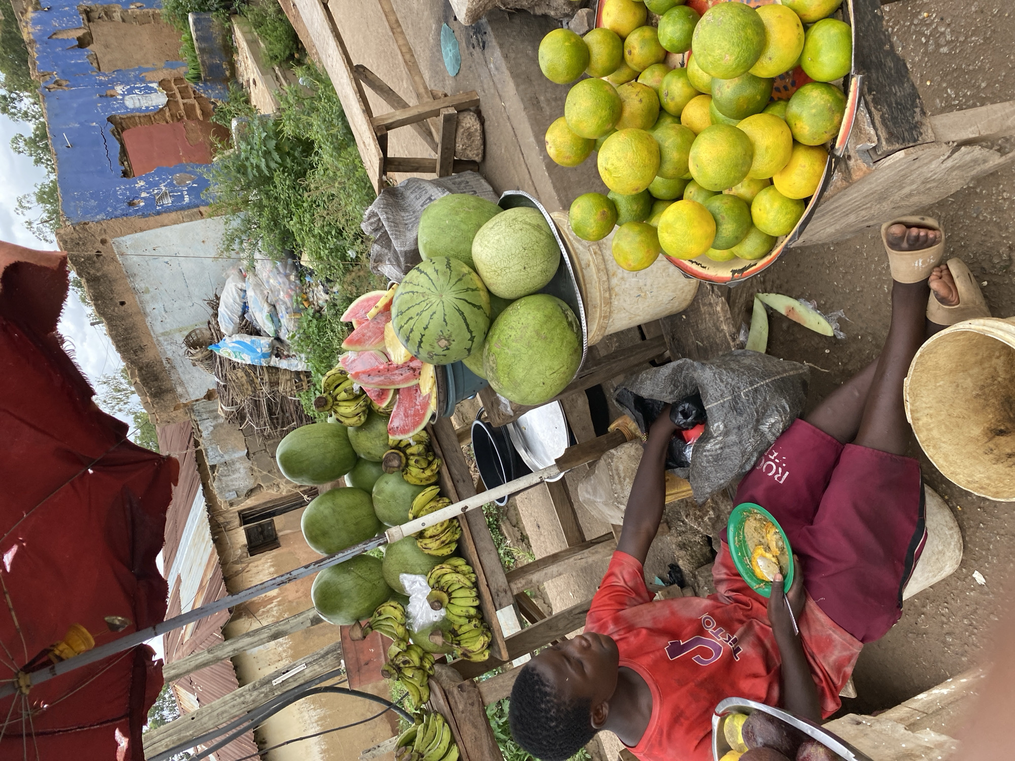 A young boy, appearing to be of Nigerian descent, is seated on a white bucket at a makeshift fruit stand under a red umbrella in Bukuru, Nigeria. He wears a red t-shirt with a "TikTok" logo and maroon shorts. The boy holds a green plate with food, appearing to be eating. The fruit stand displays an assortment of fruits, including watermelons, bananas, and oranges, arranged on wooden tables. In the background, dilapidated buildings with peeling paint and corrugated iron roofs suggest a rural or impoverished urban setting. Bags of produce and other items are scattered around the stand. The ground is dirt and gravel.