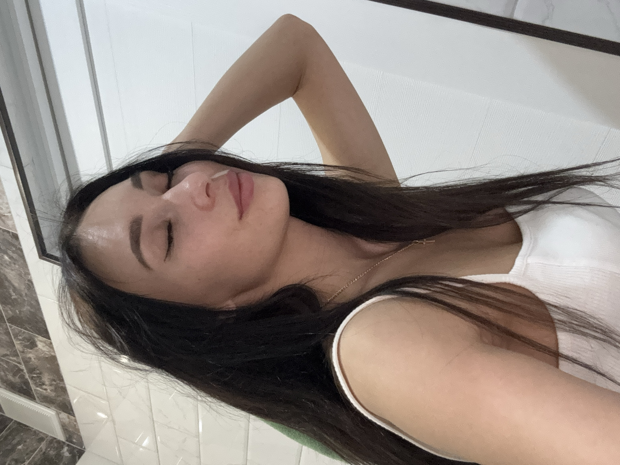 The image is a selfie of a woman with long, dark hair. She is wearing a white tank top and a gold necklace. She has her eyes closed and her arm raised behind her head. The background appears to be a bathroom with white and grey tiled walls. There is no indication of the location being Chaikovsky, Russia.