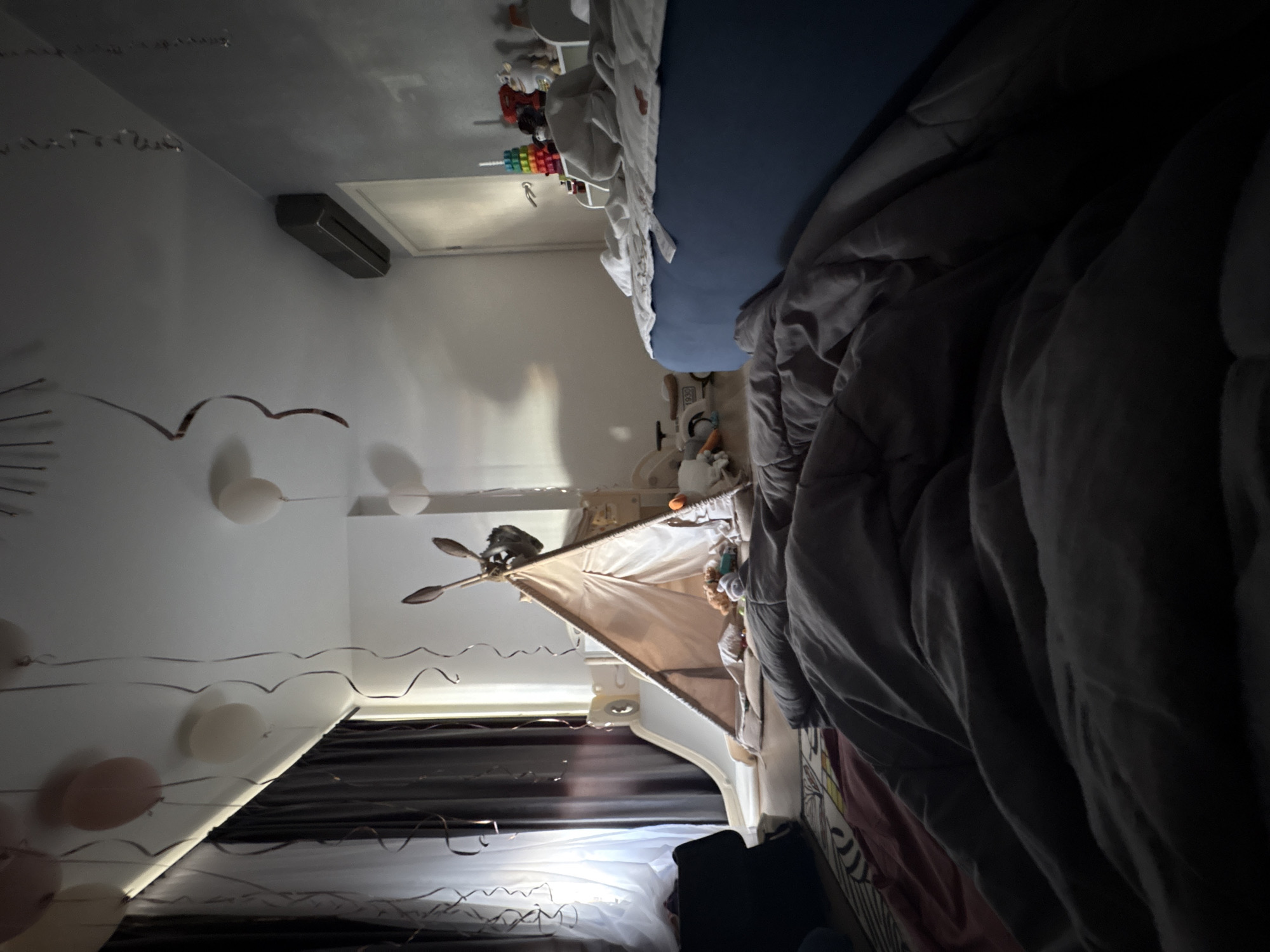 The image appears to be a skewed, low-light, cluttered interior of a bedroom. In the foreground, a portion of a bed is visible, covered with gray bedding. Balloons and streamers are hung from the ceiling. A play tent stands in the corner of the room. On the wall is an air conditioning unit. A door leading to another room is visible. There are children's toys and assorted objects on the bed. The image's skewed perspective and uneven lighting make it difficult to ascertain further details.