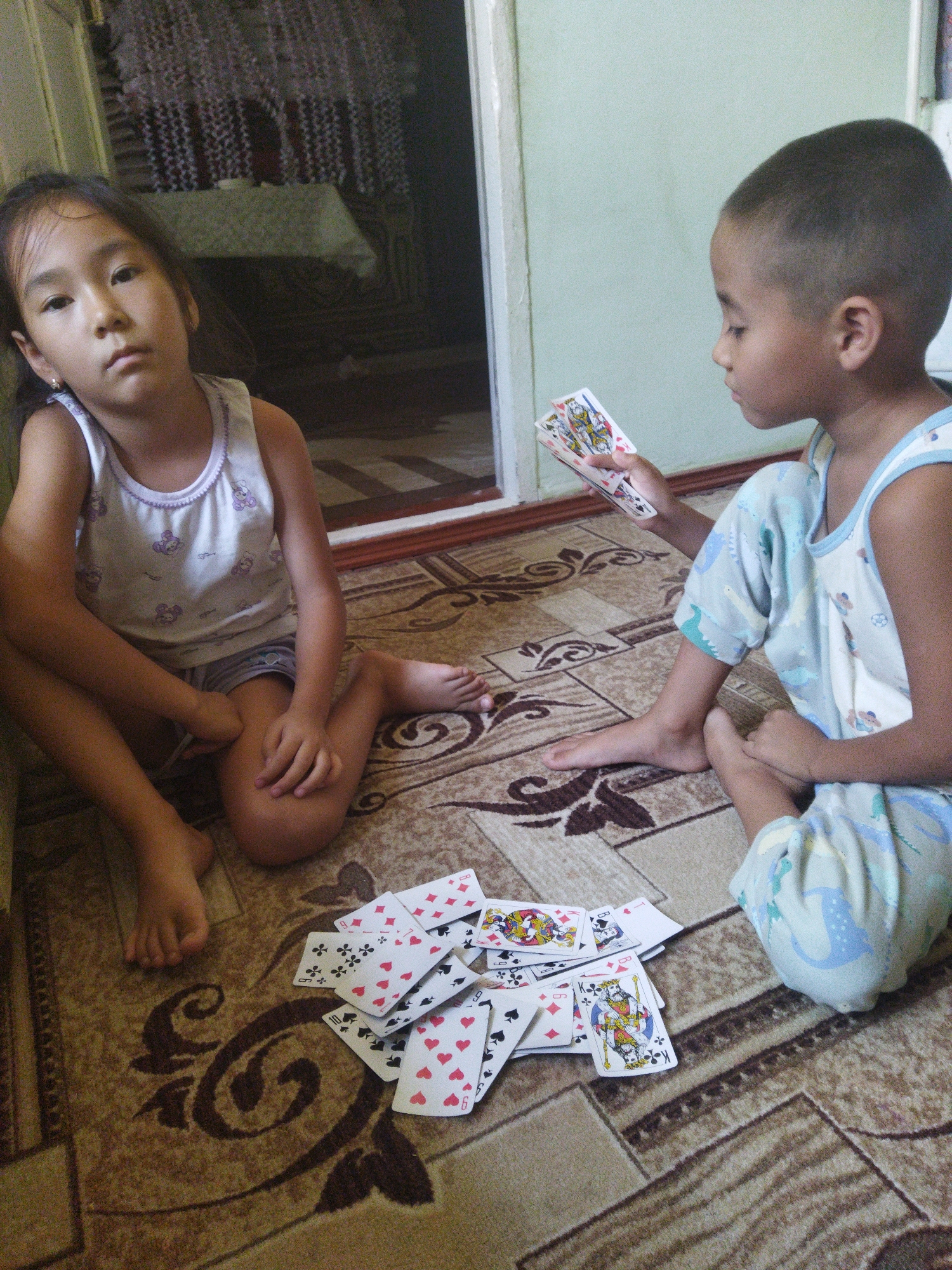 Two children, a girl and a boy, are sitting on a patterned carpet playing cards. The girl, on the left, is wearing a white tank top with purple trim and a purple pattern of small bears or bunnies. She has dark hair and is looking towards the camera with a neutral expression. She is sitting with her legs crossed and her left arm resting on her leg. The boy, on the right, is wearing a light blue tank top and pants with a light blue and green dinosaur pattern. He has short dark hair and is holding a fan of playing cards in his right hand, looking down at them. Scattered on the carpet between the children are several playing cards, some face up and some face down. The background shows a doorway with a table covered by a tablecloth in the dimly lit room beyond. The floor is covered by a beige and brown carpet with a large, dark brown swirling pattern.