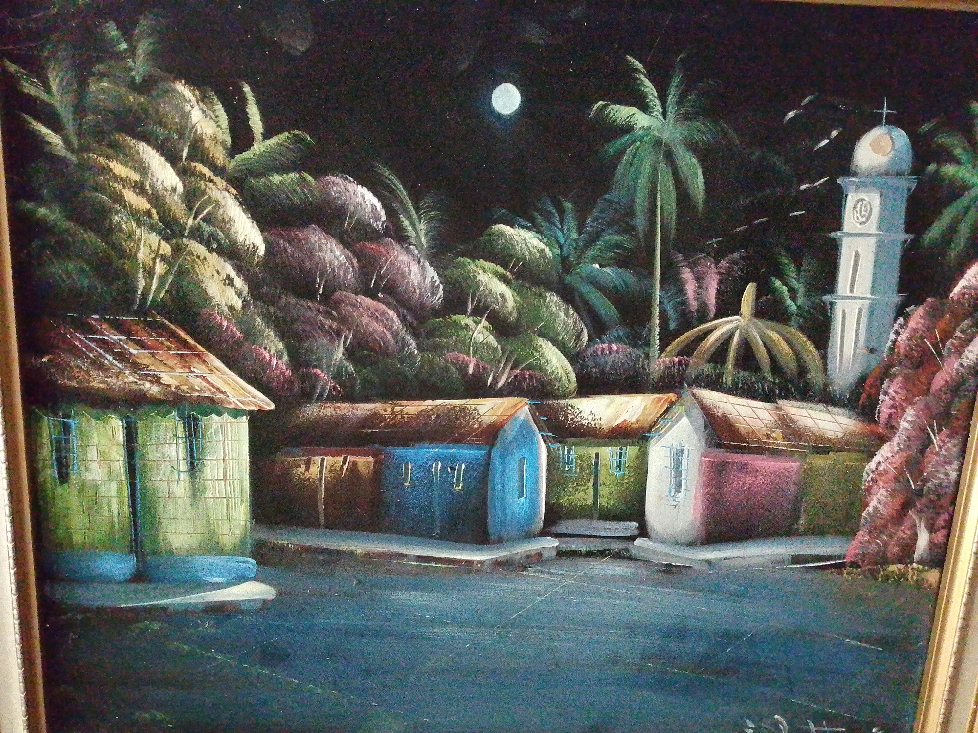 This is a painting depicting a night scene in Tinaquillo, Venezuela. Underneath a full moon, a row of four small, brightly colored houses with thatched roofs lines a waterfront. Lush vegetation and palm trees surround the houses. A tall, white church tower with a domed roof and a cross on top stands to the right of the houses. Some birds are flying around the tower. The water in front of the houses is dark and still. The painting is framed.