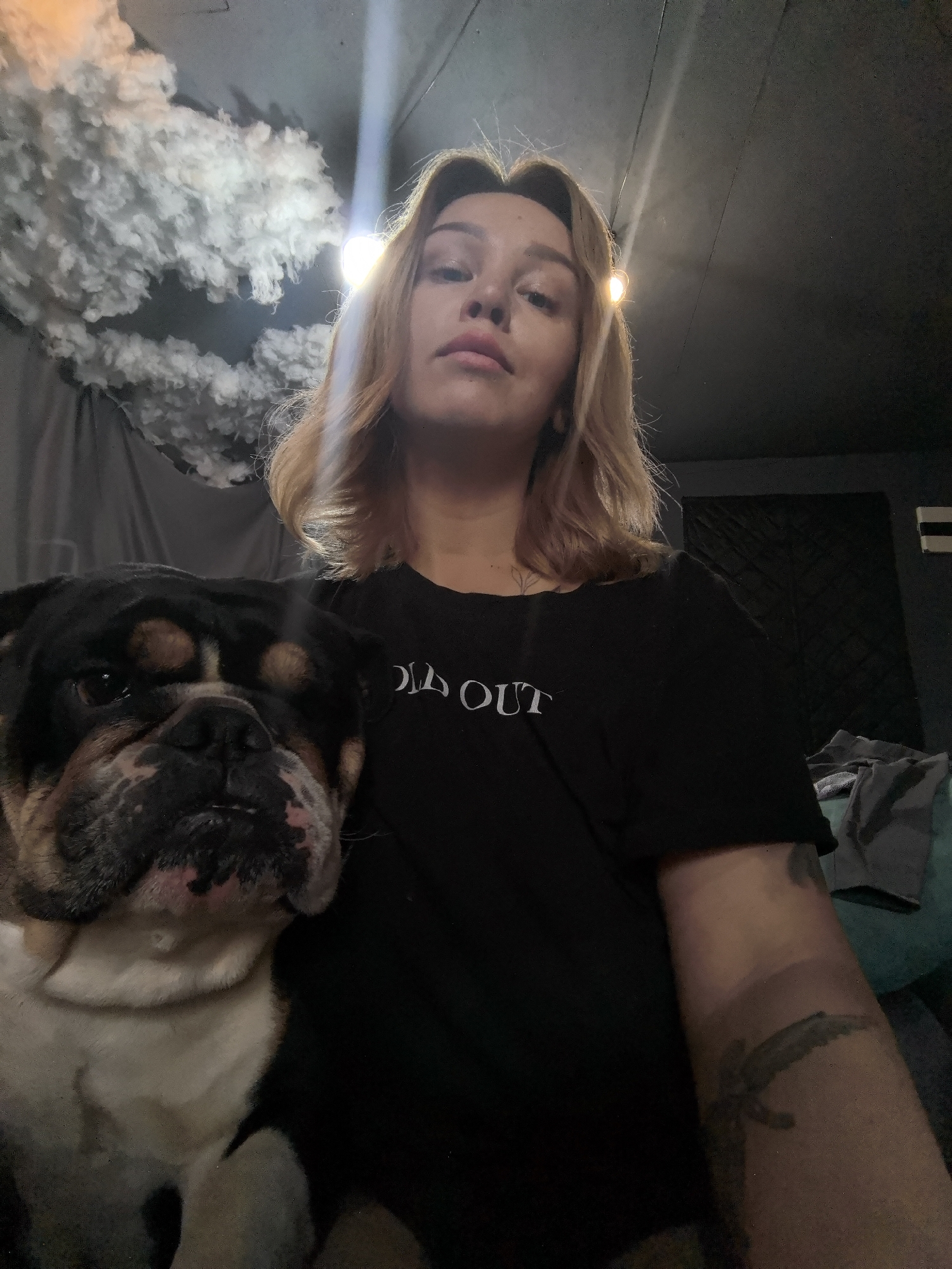 A woman with blonde hair and fair skin is in the foreground, wearing a black t-shirt with the word "SOLD OUT" printed on it. She has visible tattoos on her right forearm and neck. To her left is a bulldog with a predominantly dark face and white chest. Behind them is a dark room with a white, cloud-like structure attached to the ceiling. A light fixture is also visible on the ceiling. The location is not identifiable as Noviy, Russia.