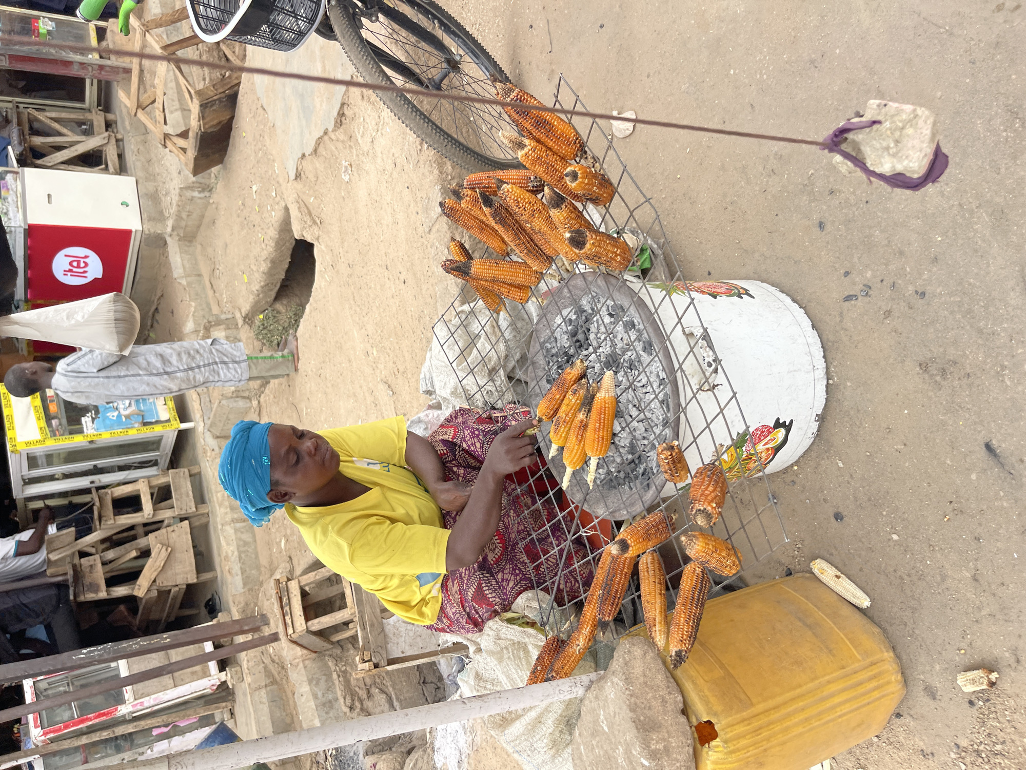 An outdoor scene captures a woman grilling and selling corn at a street stall in Chikaji, Nigeria. The central figure, a woman with dark skin, is seated on a low stool, wearing a bright yellow t-shirt and a blue headscarf. She is actively managing several corn cobs on a round metal grate placed over a charcoal fire, which appears to be housed within a repurposed, painted drum or container. Many grilled corn cobs, some showing distinct charring, are displayed on and around the grill on metal grates and white sacks, ready for customers.
To the left, a person wearing a light-colored, long-sleeved garment stands near a white and red refrigerator featuring the "itel" logo. Various wooden crates and makeshift structures are scattered throughout the background, indicative of an informal market setting. A bicycle is leaning in the upper background, and a large yellow plastic jerry can rests on the dirt ground near the corn display. The ground itself is an uneven mixture of dirt and concrete.
A notable detail in the upper right shows a wire extending across the frame with a stone tied to its end, possibly acting as a counterweight or part of a display structure. The scene is brightly lit by daylight, casting clear shadows. In the background, the word "VILLAON" is visible multiple times on what appears to be a reflective surface or product packaging. The image portrays a focused vendor engaged in her daily business.