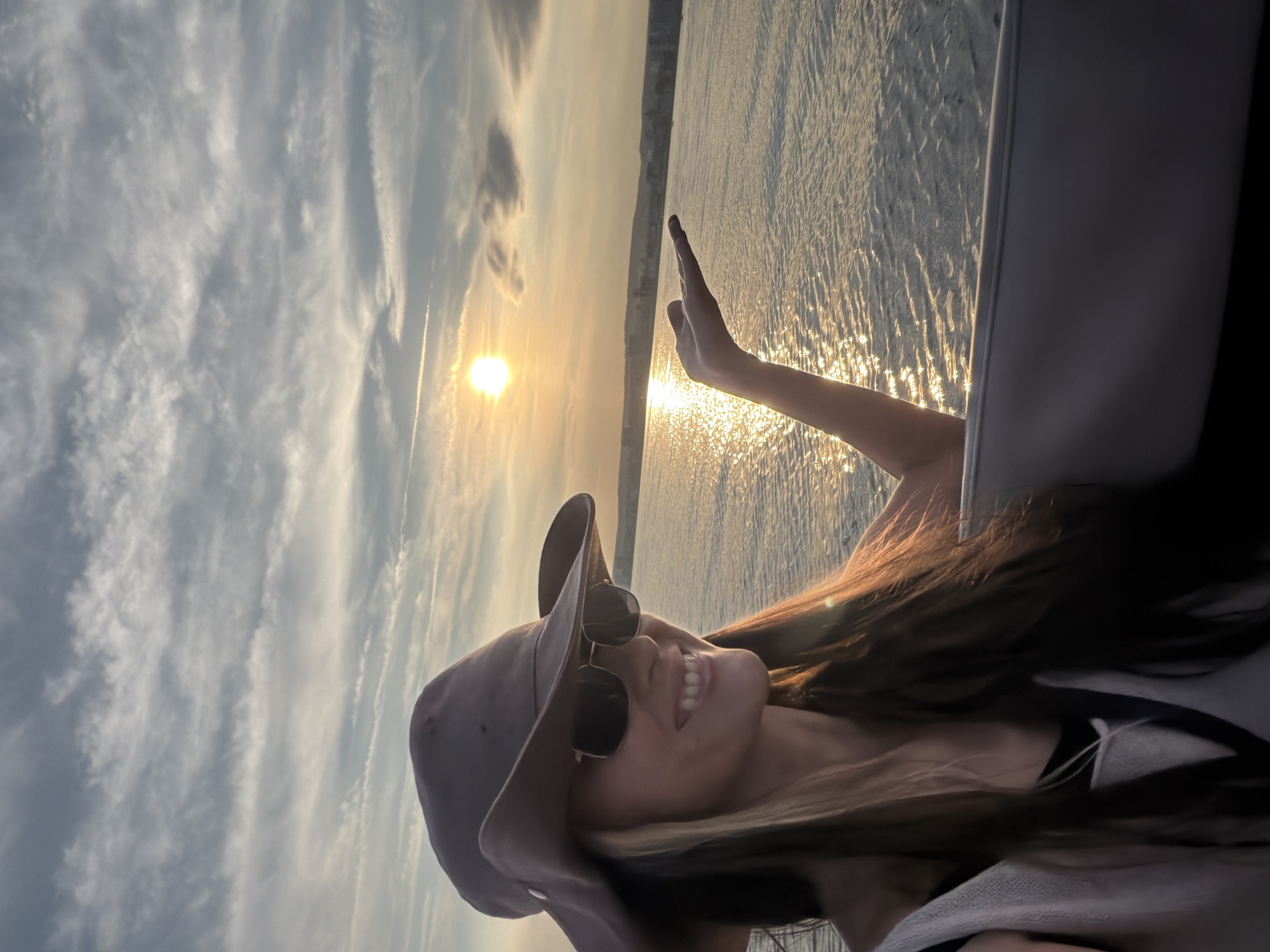A medium shot photograph captures a woman on a boat on a body of water, likely the Volga River near Engels, Russia, at sunset. The woman, wearing a wide-brimmed hat and sunglasses, smiles as she extends her left hand towards the setting sun. The sun, low in the sky, casts a golden reflection on the rippling water. In the background, a hazy cityscape is visible, hinting at the location. The sky is filled with wispy clouds, adding to the serene atmosphere. The woman is dressed in a white top over a black garment. The boat's interior, including a gray seat, is partially visible in the foreground.