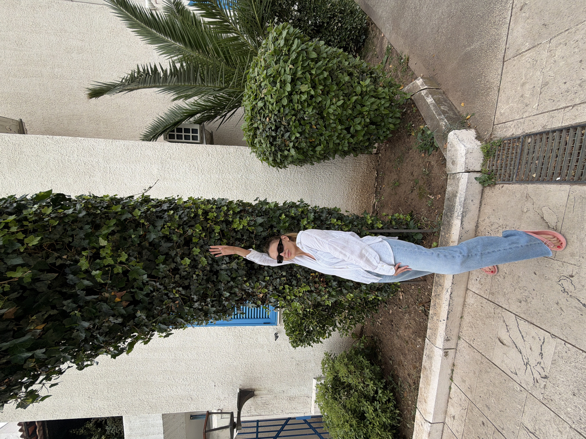 The image captures a woman posing outdoors in Budva, Montenegro, set against a backdrop of white architecture and dense greenery. The scene is brightly illuminated, indicating daytime with soft, possibly overcast, lighting.
The central subject is a woman with light hair, seen lying horizontally against an ivy-covered wall or bush. She wears a casual outfit consisting of a white long-sleeved collared shirt, light blue denim jeans, and pink flip-flops. Dark sunglasses adorn her face, and she has a relaxed expression while looking towards the camera. Her right arm is extended upwards and bent slightly at the elbow, adding to her casual pose.
Behind the woman, a white, textured building forms a significant part of the background. It features a prominent blue-shuttered window to the left and a smaller, gridded window or vent to the right. Lush vegetation surrounds the building, including a tall palm tree extending its fronds into the upper right corner of the frame, and several rounded, dense green bushes. The ivy wall the woman leans against is thick and dark green.
The ground consists of light gray, rectangular paved stones with visible dark vein patterns, typical of natural stone. A rectangular metal drainage grate is embedded in the pavement on the right. Alongside the paved area, there are concrete curbs and patches of earth with sparse green plants. The overall setting suggests a pleasant, Mediterranean-style outdoor environment, possibly a garden path or a street alongside a building. No visible text is present in the image.