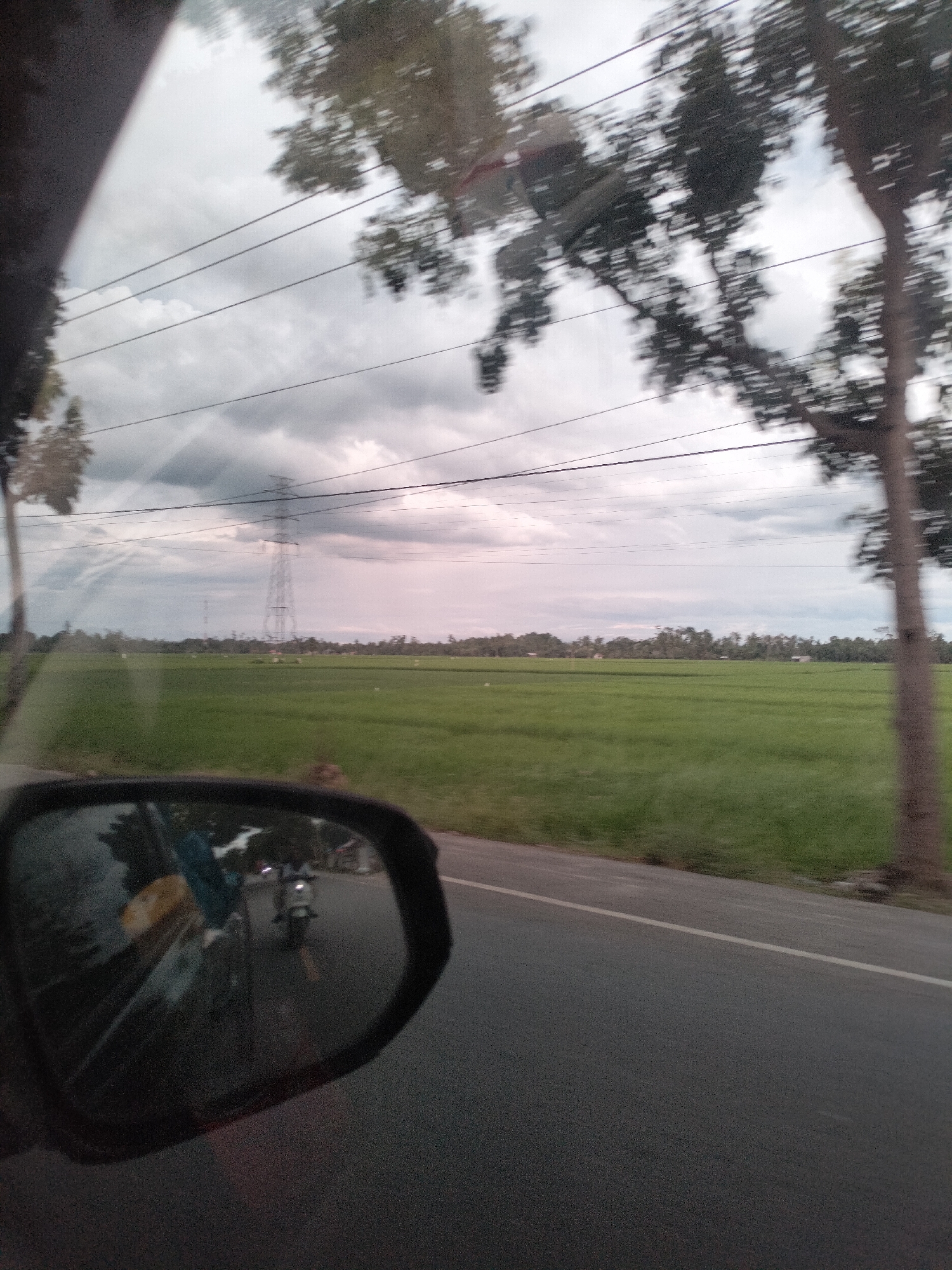 The image captures a view from a vehicle traveling along a road in the vicinity of Putoh Sa, Indonesia. A side-view mirror on the left side of the frame reflects a motorcycle with a rider, and part of the vehicle's interior, including a yellow object and a blue garment. Outside the vehicle, the scene unfolds with a wide, green rice paddy stretching towards a treeline in the distance. A tall, lattice electricity pylon is visible in the middle ground, set against a cloudy sky with power lines strung across it. Trees line the right side of the road, with foliage blurring due to motion. The road itself is asphalt with a white line marking its edge.