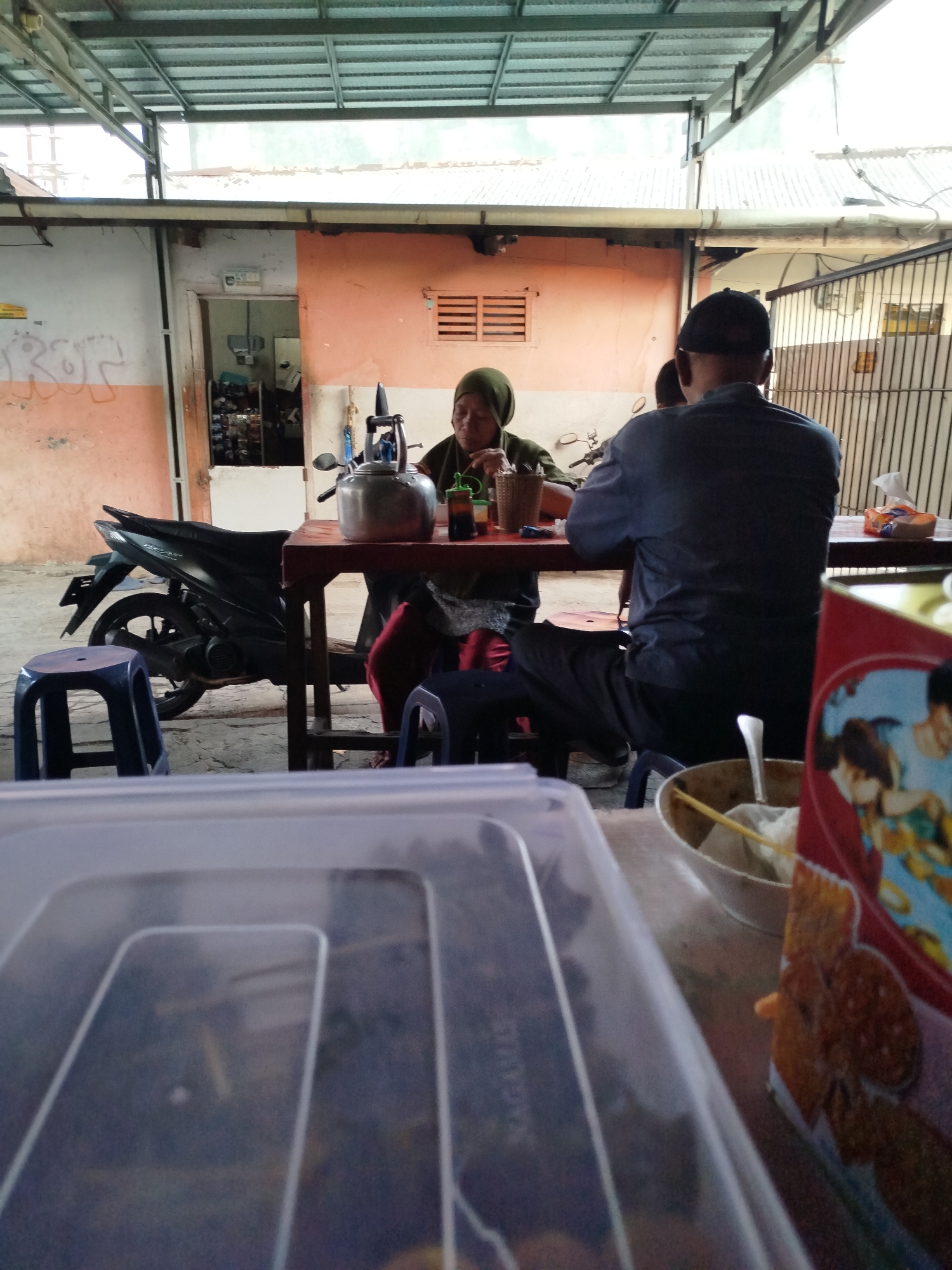 The image depicts an outdoor eating area, likely a food stall or a simple restaurant, in Depok, Indonesia. In the foreground, a blurred view of plastic food containers dominates the lower half of the frame. The middle ground reveals a wooden table set with two people. A woman wearing a hijab and an olive-green shirt is seated, appearing to eat. In front of her is a metal kettle and a woven basket. To her right, a man in a dark blue shirt and a black cap is seated, facing away from the camera. He is seated on a blue plastic stool. Behind the table, a black motorcycle is parked. The background shows a building with an orange-tinted wall and a metal roof structure. The overall scene suggests a casual dining experience in a modest urban setting.