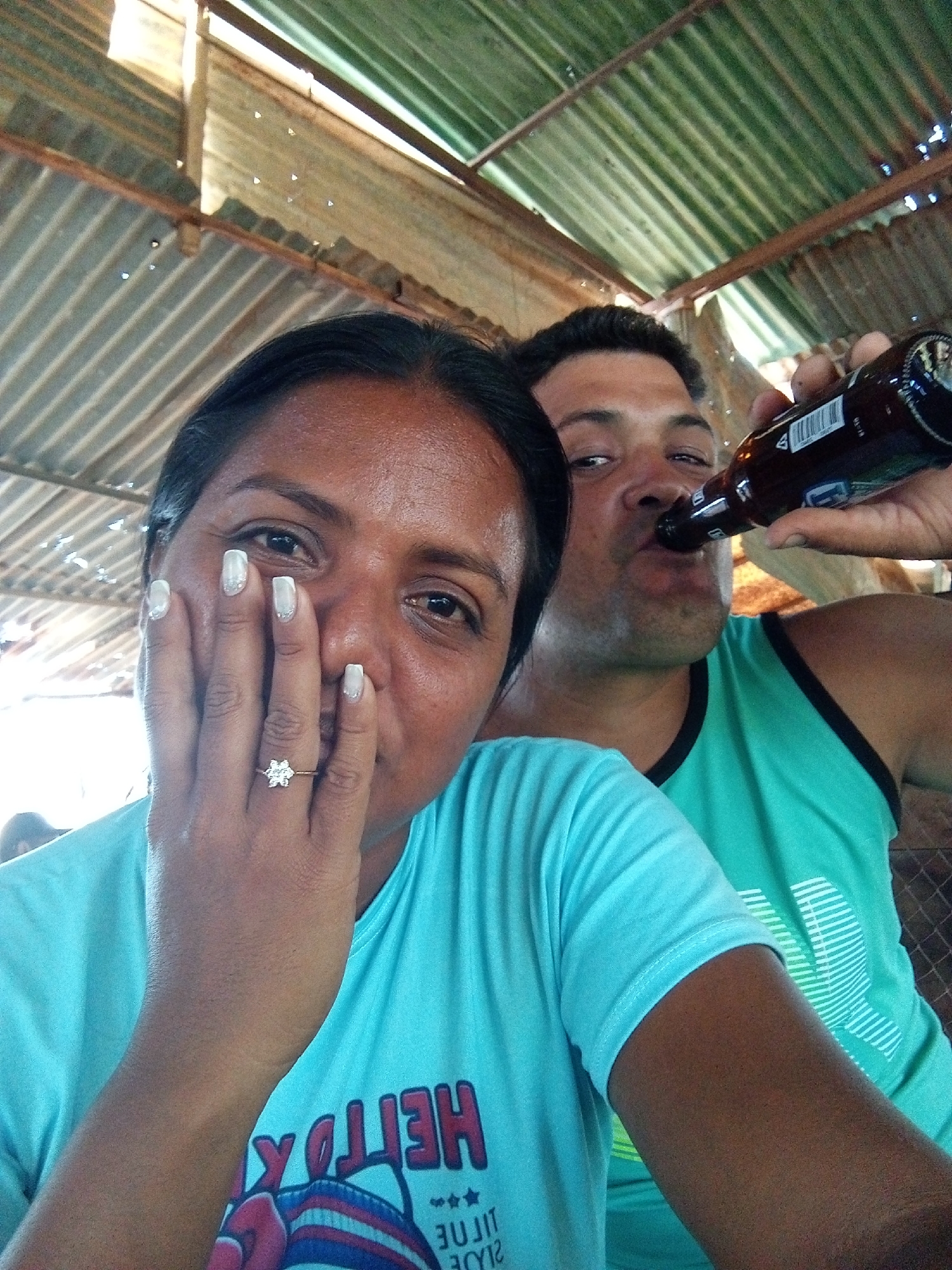 Two adults are indoors, likely in Unknown, Unknown, based on the appearance of the surroundings. A woman with dark hair wears a light blue shirt and a floral-shaped ring on her left ring finger. She partially covers her face with her hand. A man behind her, wearing a green tank top, drinks from a brown glass bottle. The setting features corrugated metal roofing.