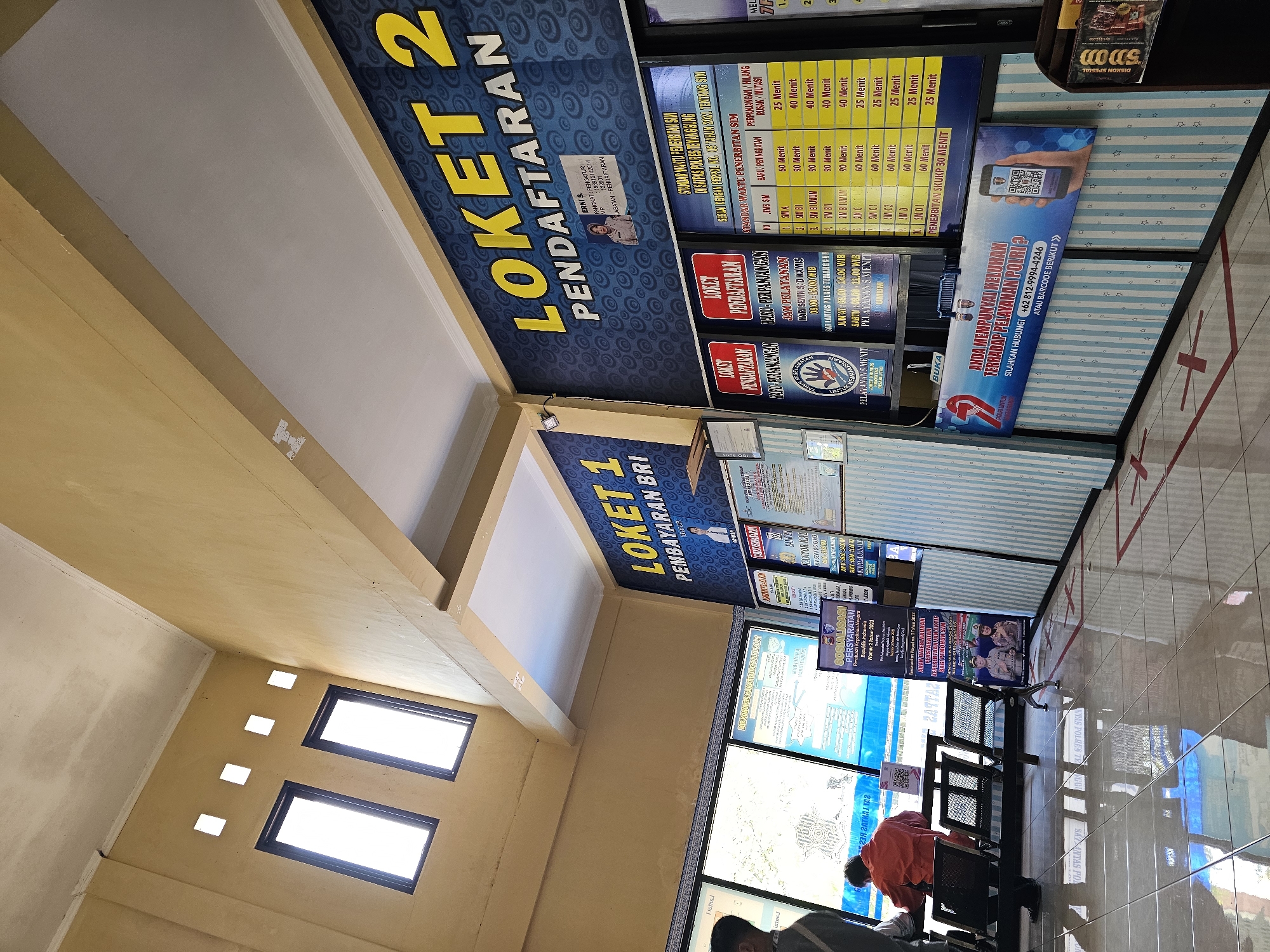 The image shows the interior of a police station service counter, likely the Satpas (Surat Izin Mengemudi - Driver's License) office. The prominent signage indicates "LOKET 1 PEMBAYARAN BRI" (Counter 1 BRI Payment) and "LOKET PENDAFTARAN" (Registration Counter) with the number 2. The walls are decorated with a blue and white striped pattern and official posters.
Several service information posters are visible, detailing service hours and requirements for SIM (Surat Izin Mengemudi) applications. For example, one poster lists "STANDAR WAKTU PENERBITAN SIM" (Standard SIM Issuance Time) with categories like SIM A, SIM B1, SIM B2, SIM C, SIM D, and their respective processing times for new applications and renewals. Another poster prominently features "ANDA MEMPUNYAI KELUHAN TERHADAP PELAYANAN POLRI?" (Do you have complaints about Police Services?) with a contact number and a QR code.
In the foreground, the tiled floor reflects the lighting and signage. There are several metal chairs with black seats arranged in rows. A person in an orange shirt is seated at a desk, facing away from the camera, likely interacting with a service window. Another person, partially visible on the left, appears to be standing. The overall atmosphere suggests a functional government service area. The presence of "SATUAN LALU LINTAS POLRES TEMANGGUNG" (Traffic Unit Temanggung Polres) on a poster indicates the location as Temanggung, Indonesia, specifically within the Madureso sub-district of Temanggung Regency.