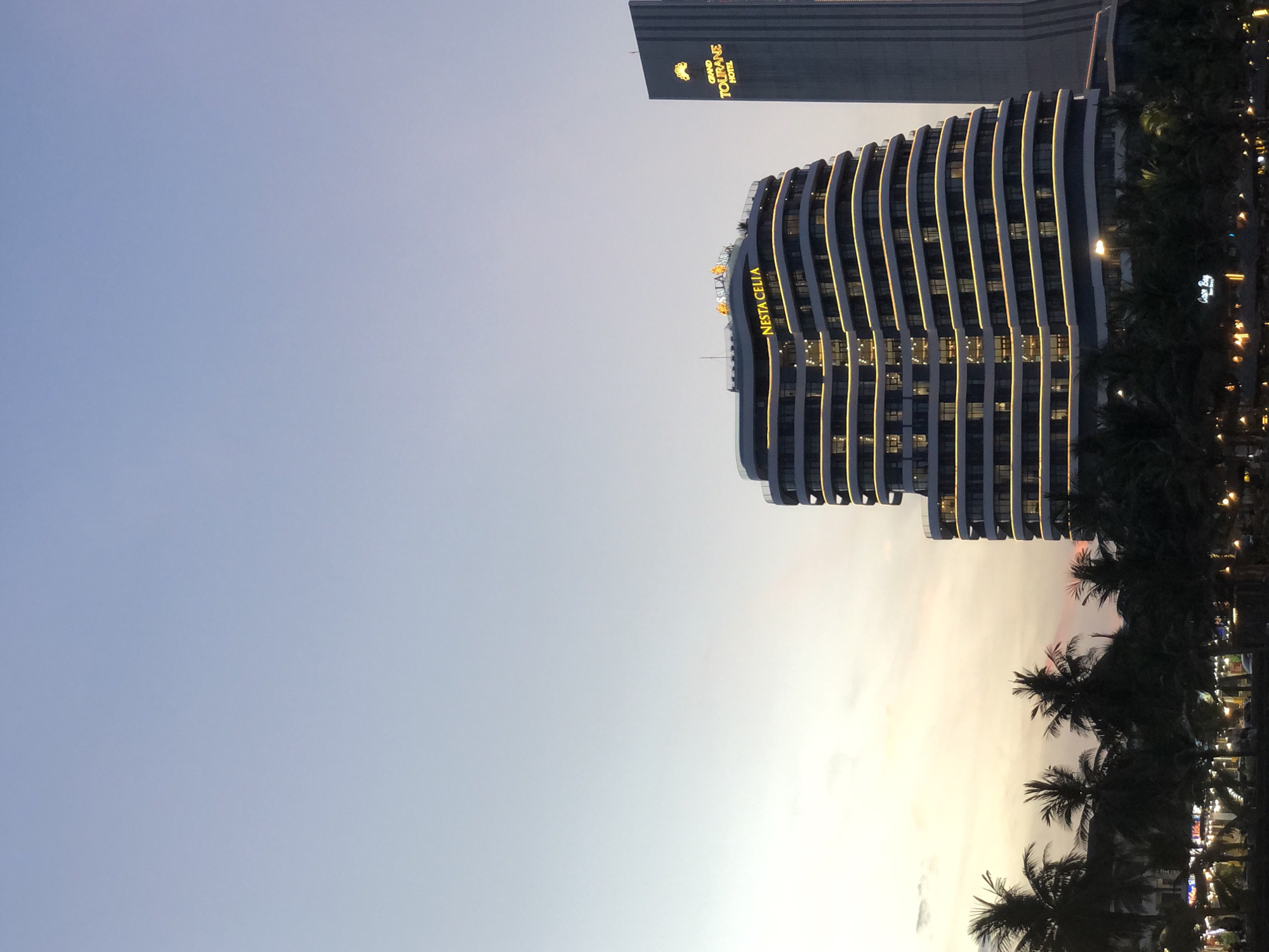This image captures a twilight view of the beach and coastline in Da Nang, Vietnam, with two prominent buildings illuminated by evening lights. The left building, with its distinctive curved, layered facade, is labeled "NESTA CELIA" in bright yellow lettering. To the right, a taller, more rectangular building displays the prominent sign "GRAND TOURANE HOTEL" also in yellow, accompanied by a golden graphic. In the foreground, silhouetted palm trees line the beachfront, with the glow of lights from what appears to be a promenade or resort area visible beneath them. The sky is a gradient of pale blue and soft orange, indicative of sunset or sunrise.