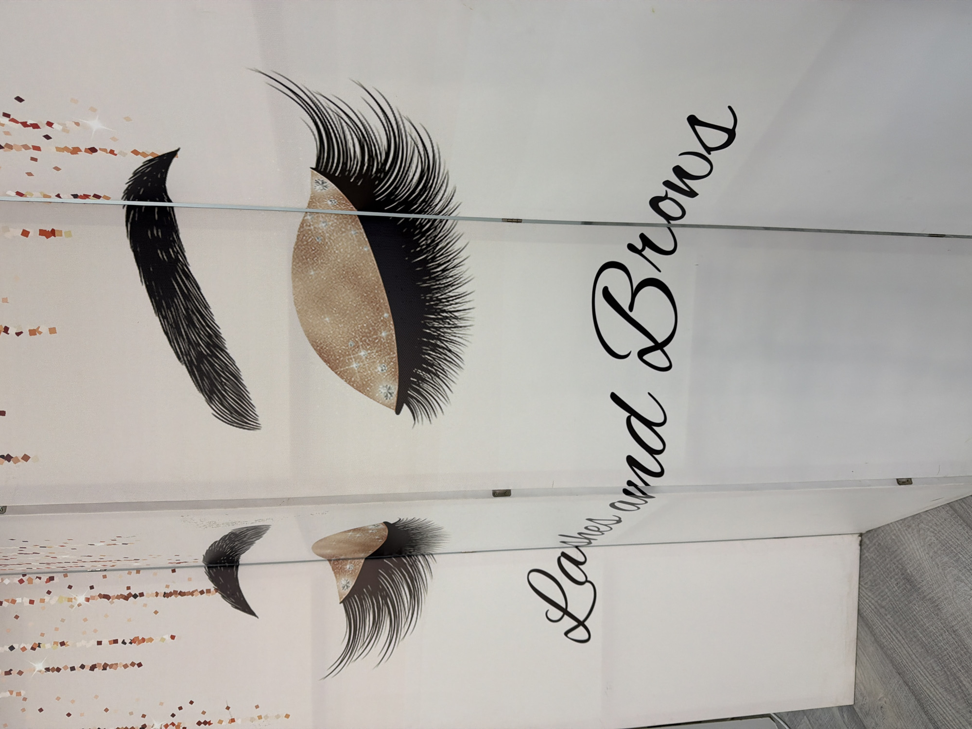 This is a close-up, eye-level, interior shot of a white room, likely a beauty salon, with a decorative partition or screen. The partition features an artistic design of stylized, closed eyes with long eyelashes and glittery eyeshadow in gold and bronze tones. Above the eyes, there are decorative strands of confetti or glitter in gold and rose gold hues. Below the eyes, the words "Lashes and Brows" are written in a black, cursive font. The partition appears to be made of panels with vertical seams and possibly glass elements. The floor is a light gray wood-grain laminate. The overall impression is one of a clean, modern, and feminine aesthetic, suggestive of a spa or salon environment. The scene is static and does not depict any action or people. There is no information to suggest this is in Kyiv, Ukraine.