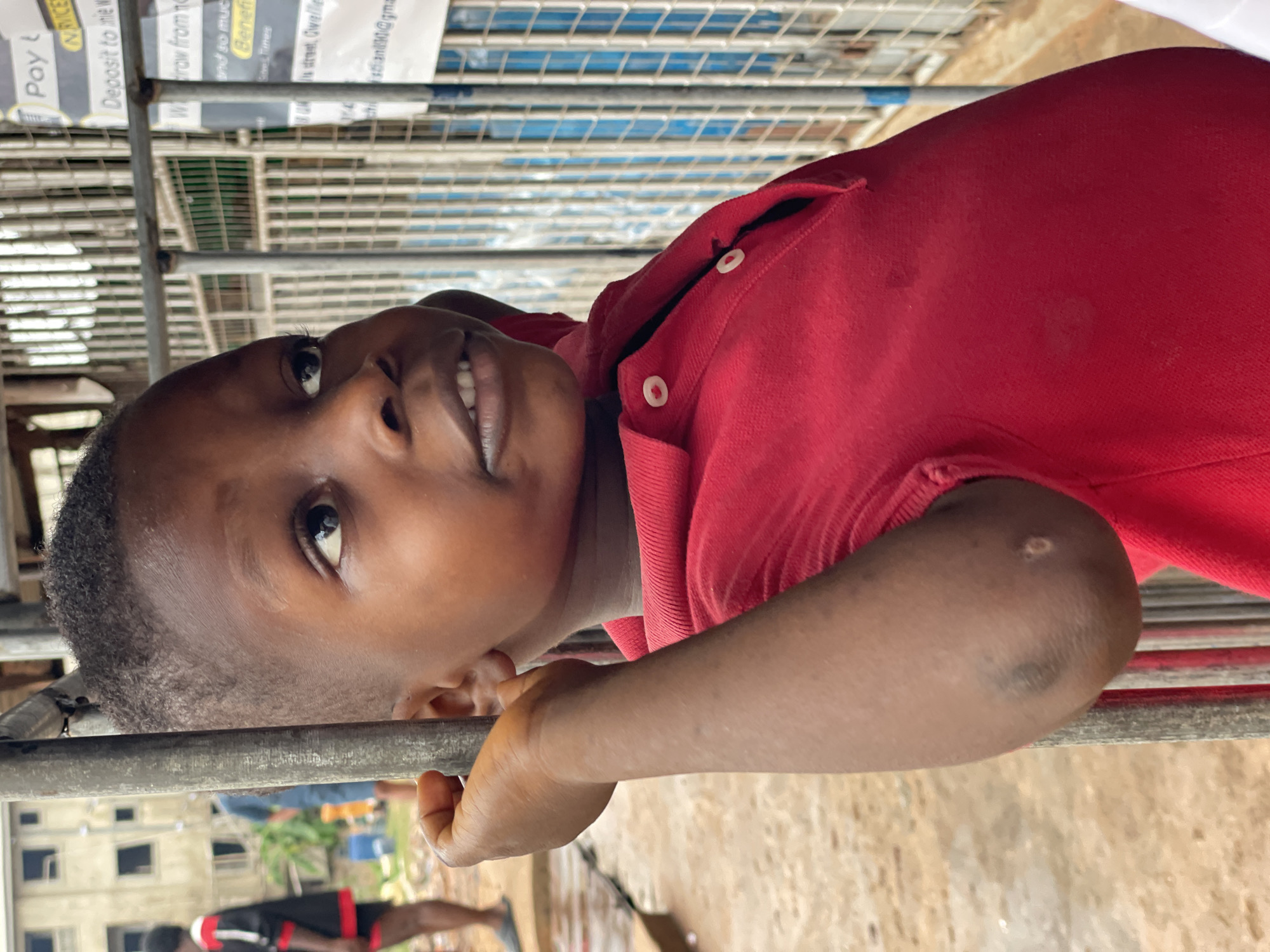 A young boy, likely in Onitsha, Nigeria, is pictured in an outdoor setting. He is wearing a red polo shirt and leaning on a metal railing. In the background, there's a building and a person walking. The boy is looking upwards, possibly towards the camera. A sign containing business information is visible in the background. The text on the sign contains the words "Pay", "IN RICE", "Deposit", "Widraw", "Benefi", "Gree Times", and the contact email "christian90@gmail.com". The sign suggests the location is near UK, Owelle in the city of Onitsha.