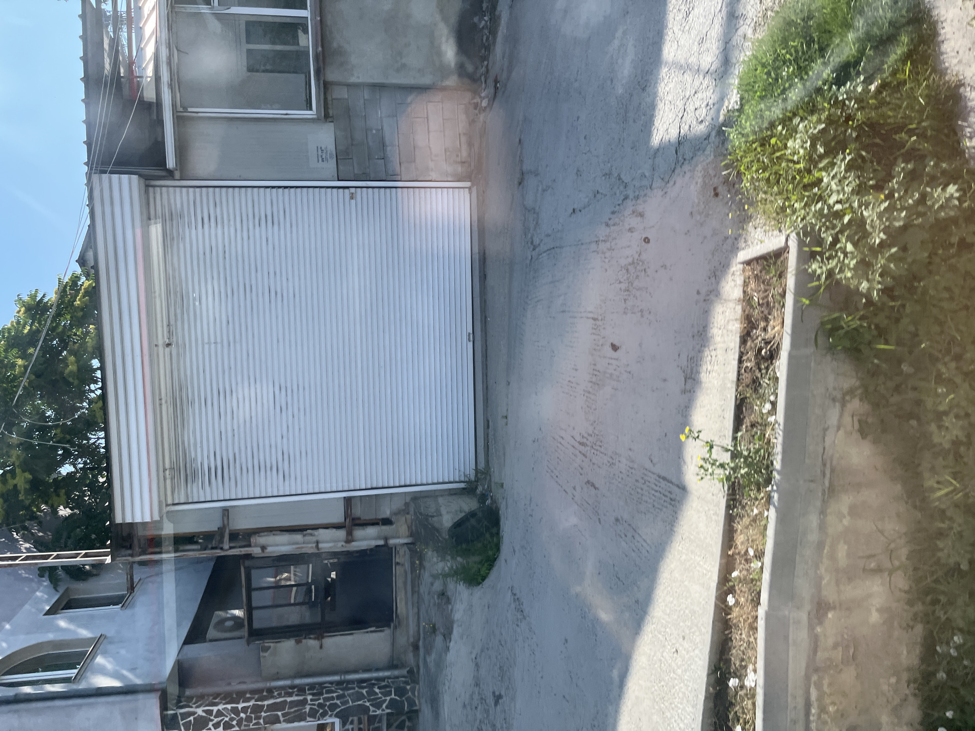 The image captures an outdoor scene in Varna, Bulgaria, featuring a commercial or industrial building with a large, white, corrugated metal roll-up door dominating the center. To the left of the door is a dark doorway with a metal frame and what appears to be an air conditioning unit above it. Adjacent to this, on the far left, is a building facade with a patterned stone design and an arched window. To the right of the large roll-up door, a window with a white frame is visible on a concrete block wall. The ground in front of the building is paved with concrete, showing some wear and tear, and slopes downwards towards the viewer. A concrete planter box runs along the bottom of the frame, containing dry grass and a few small yellow and white flowers, with a dense patch of green vegetation to its right. The sky above is clear and blue. The scene is illuminated by bright sunlight, casting shadows on the ground. There are no people or animals visible in the image.