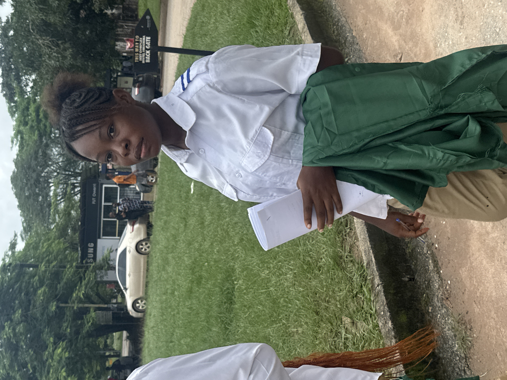 The image depicts a young woman standing in front of a building that appears to be part of a campus in Ihiagwa, Nigeria. She is wearing a white shirt with blue stripes on the shoulders and tan pants. She is holding a white document and a folded green cloth. In the background, there are trees, a car, and a signpost that reads "THIS WAY TO HOSTEL COMPLEX BACK GATE". Other people are visible in the background, some standing near a building with "SUNG" written on it, and others near cars. The ground in the foreground is a dirt path with some debris. The overall lighting suggests an overcast day.