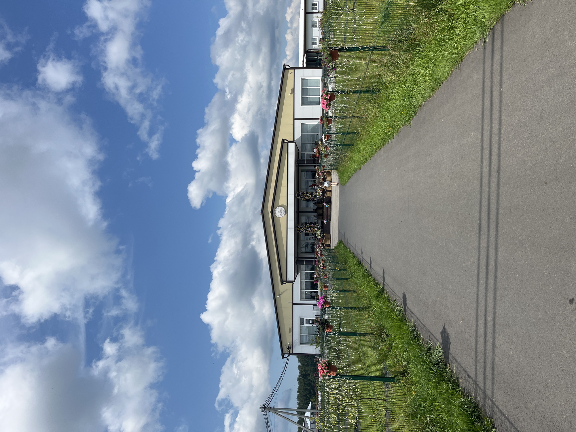 The image depicts a frontal view of a long, modern building with a light yellow roof and white walls, situated in a rural environment. A paved walkway leads directly towards the entrance of the building. The building appears to be a commercial establishment, possibly a greenhouse or a market, given the abundant flowers in pots lining a green fence on either side of the walkway.
The environment is characterized by a bright, sunny day with a clear blue sky dotted with fluffy white clouds. To the left of the building, a utility pole with wires is visible, suggesting proximity to infrastructure. Lush green grass and vegetation grow along the fence and surrounding areas.
There are a few people visible. One person, wearing a white dress, is walking away from the camera towards the building. Inside the building's entrance area, several other figures can be seen, partially obscured by decorative floral displays. The overall scene suggests a well-maintained, accessible location, likely a place of business or public attraction in the village of Akhlebaikha (Kovriginskii S/S), Russia.