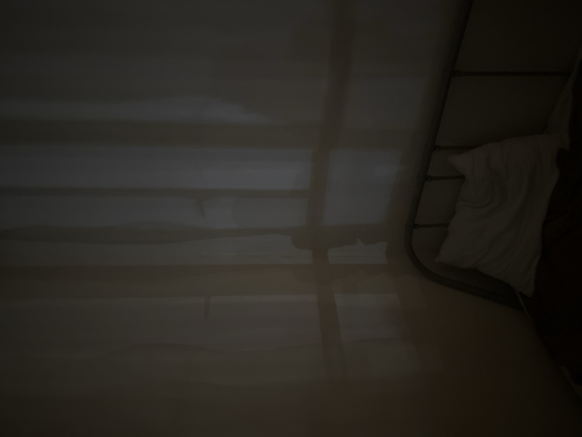 The image depicts a dimly lit bedroom interior, possibly in Sinyavino, Russia, with a stark and unsettling ambiance. A metal-framed bed with a white pillow and dark bedding occupies the lower right portion of the frame. The walls are a pale, neutral color, illuminated by faint light filtering through what appears to be a window covered by sheer curtains. The light creates dramatic shadows on the wall, with vertical lines from the curtains and a larger, indistinct shadow that could be interpreted as a human form. The overall impression is one of quietude and potential unease, with the shadows suggesting a presence or a lingering past. There are no visible people, and the scene is static.