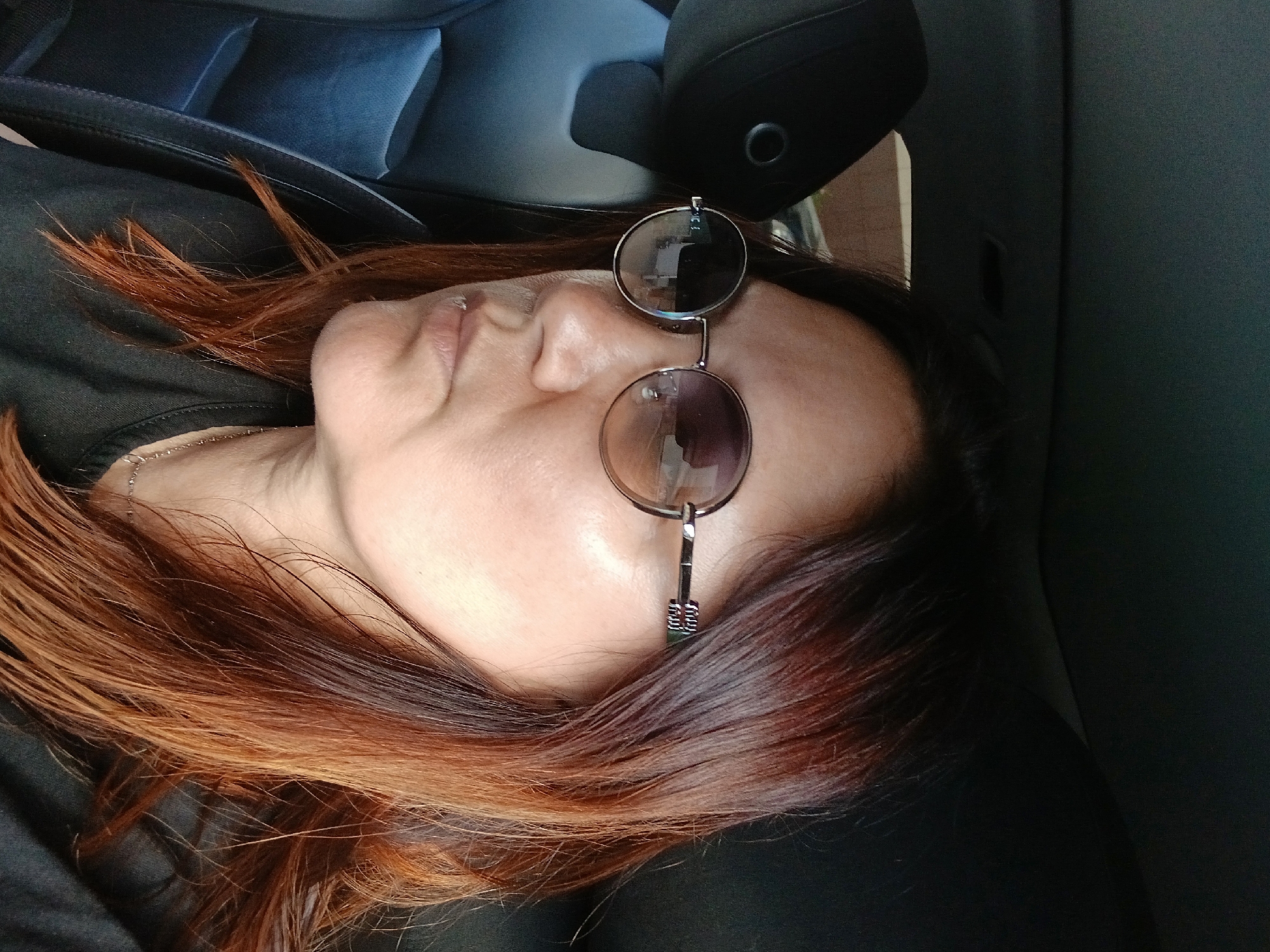The media file is a close-up, slightly tilted photograph of a person wearing round-lensed sunglasses. The subject is lying down, possibly in a vehicle, with their head resting on a dark surface. Their eyes are closed, and their mouth is slightly open, suggesting they are sleeping or resting. Long, reddish-brown hair is spread around their head and shoulders. A thin silver necklace is visible on their collarbone. The sunglasses have thin metal frames and appear to be tinted. The background consists of dark upholstery, possibly from a car seat, with some lighter blue-grey cushioning visible behind the subject's head. The lighting suggests it could be daytime, but the exact time and weather conditions are not discernible. There is no visible text in the image.